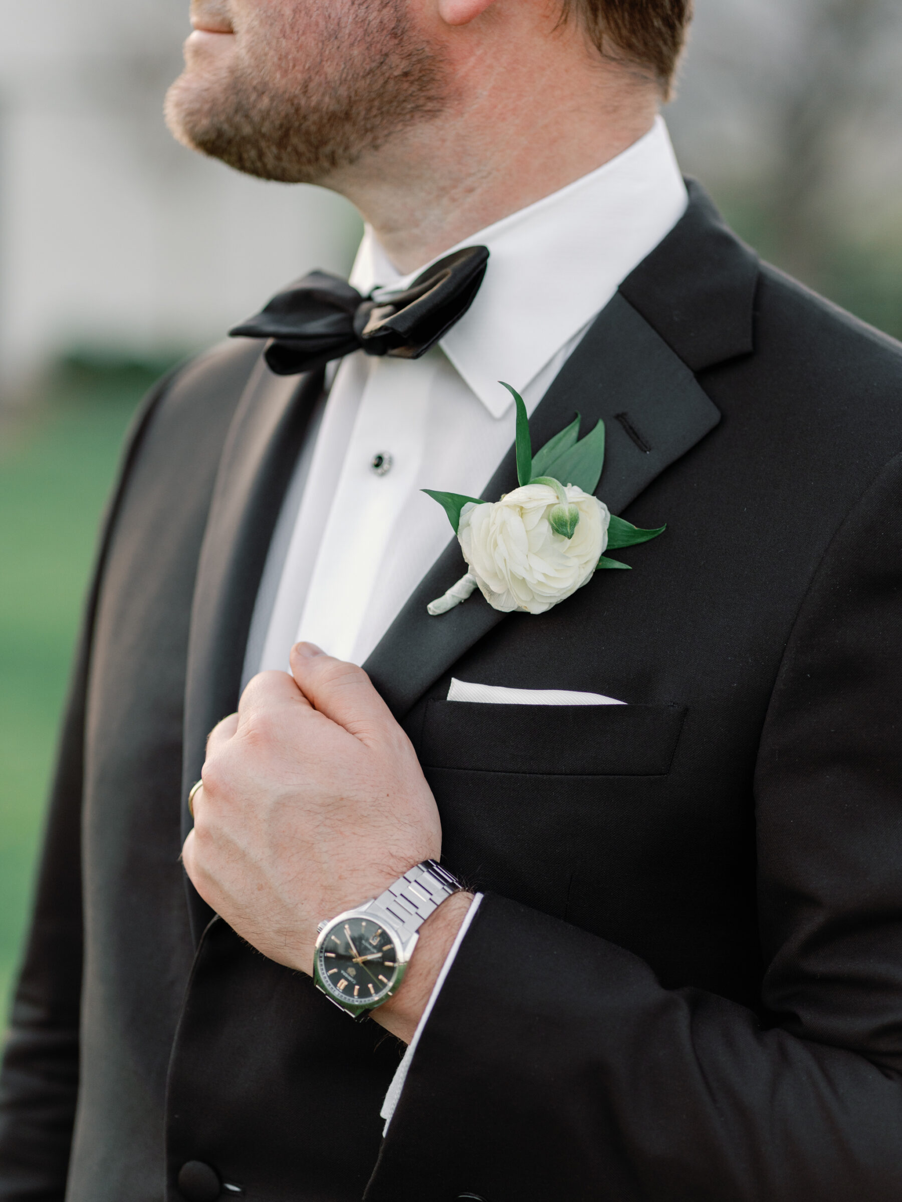 A close-up of a groom in his tuxedo, adjusting his jacket with his hand. The groom's boutonnière, a white ranunculus flower with greenery, is pinned to his lapel. His wrist is adorned with a silver watch, adding a touch of elegance. The soft focus on the background highlights the groom’s refined, classic style.