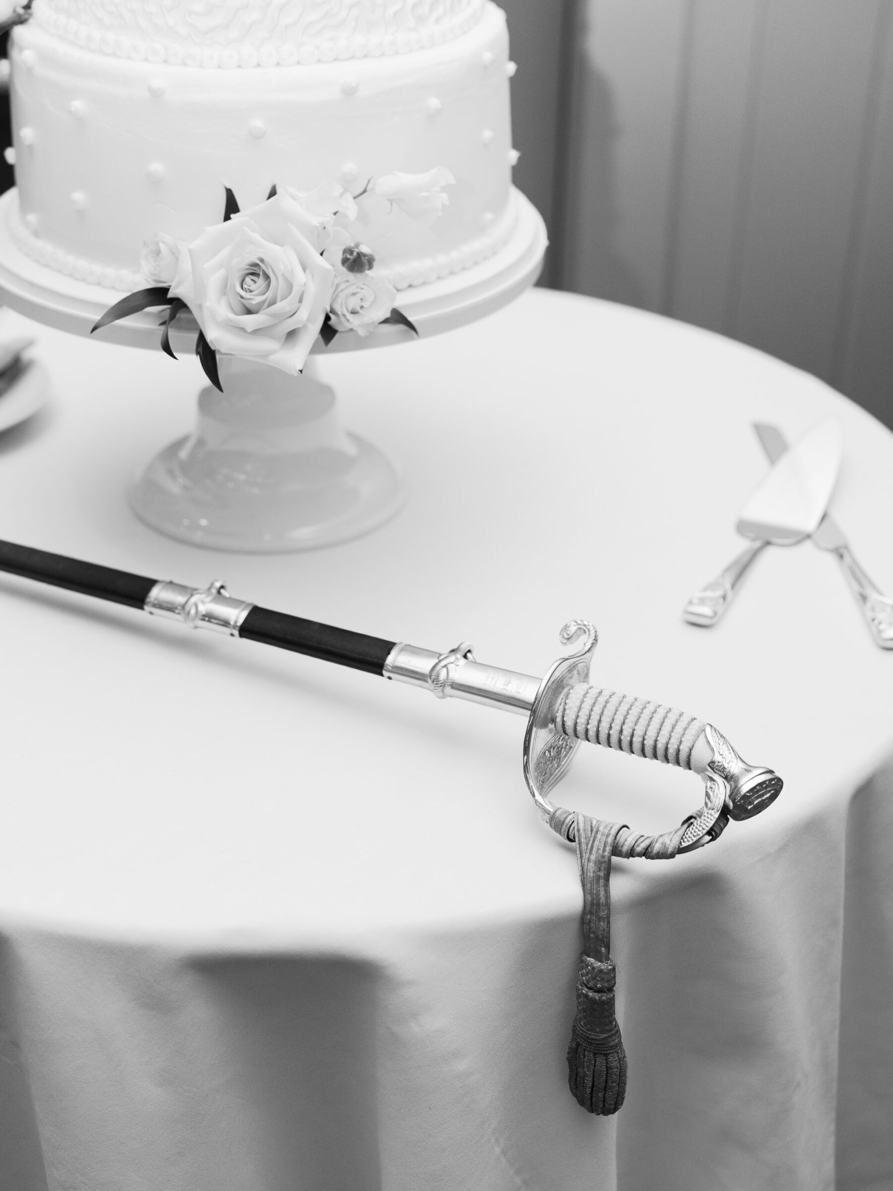 A close-up black-and-white photo of a wedding cake with soft pink roses and delicate pearl detailing, sitting on a pedestal table. A ceremonial sword rests beside the cake, its handle wrapped in intricate cord and adorned with a silver guard, symbolizing a regal touch. Cake-cutting utensils lie on the table near the sword, creating a refined and elegant setting.