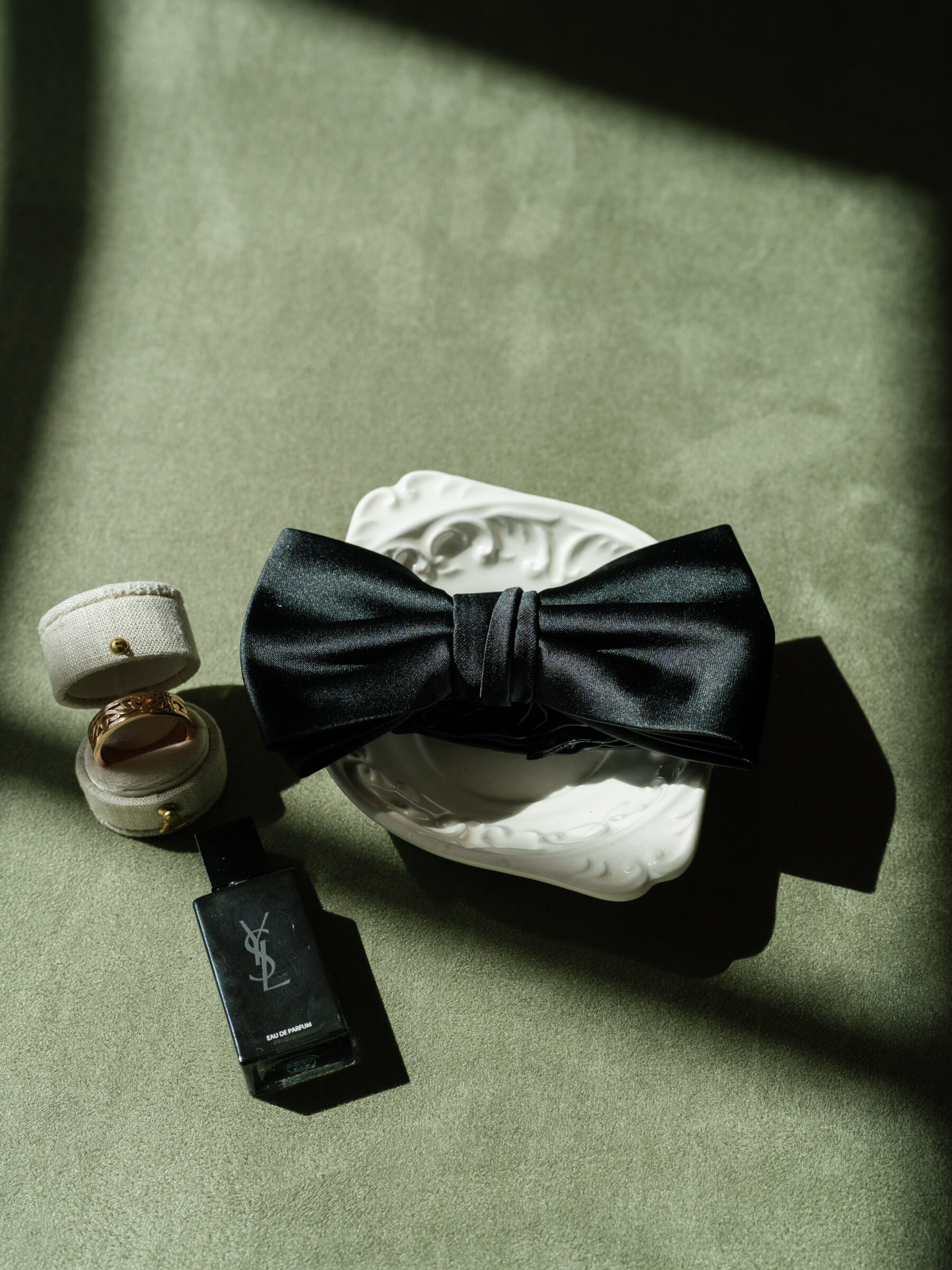 A close-up shot of a groom's wedding details featuring a black satin bow tie, a bottle of Yves Saint Laurent perfume, and two wedding rings placed elegantly on a white dish, all set against a soft green velvet backdrop. The light highlights the luxurious textures and details of the items.