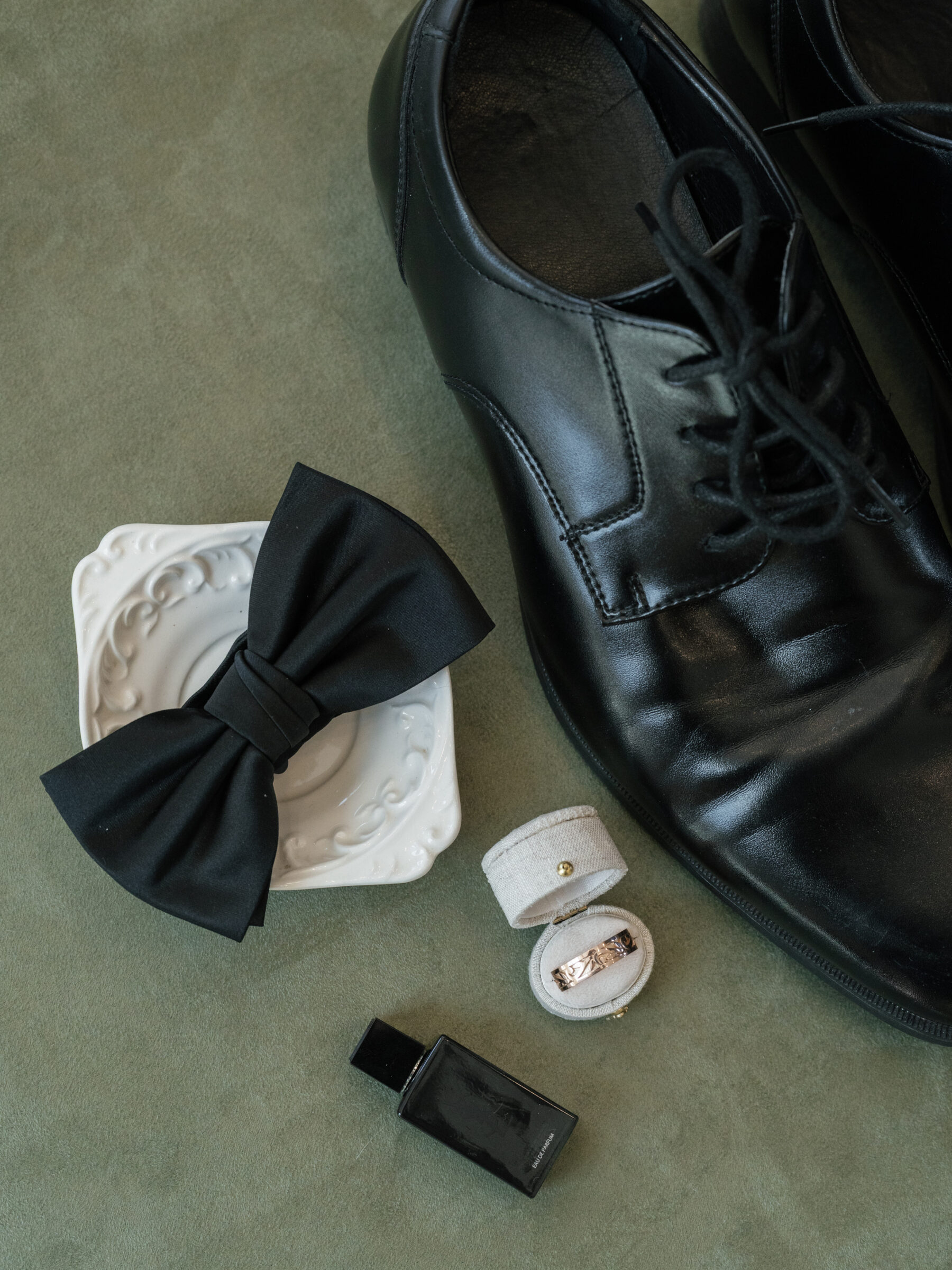 A flat lay of a groom's wedding essentials: a black bow tie resting on a small white dish, black leather dress shoes, a ring box holding a gold wedding band, and a bottle of Yves Saint Laurent cologne placed on a soft green surface.