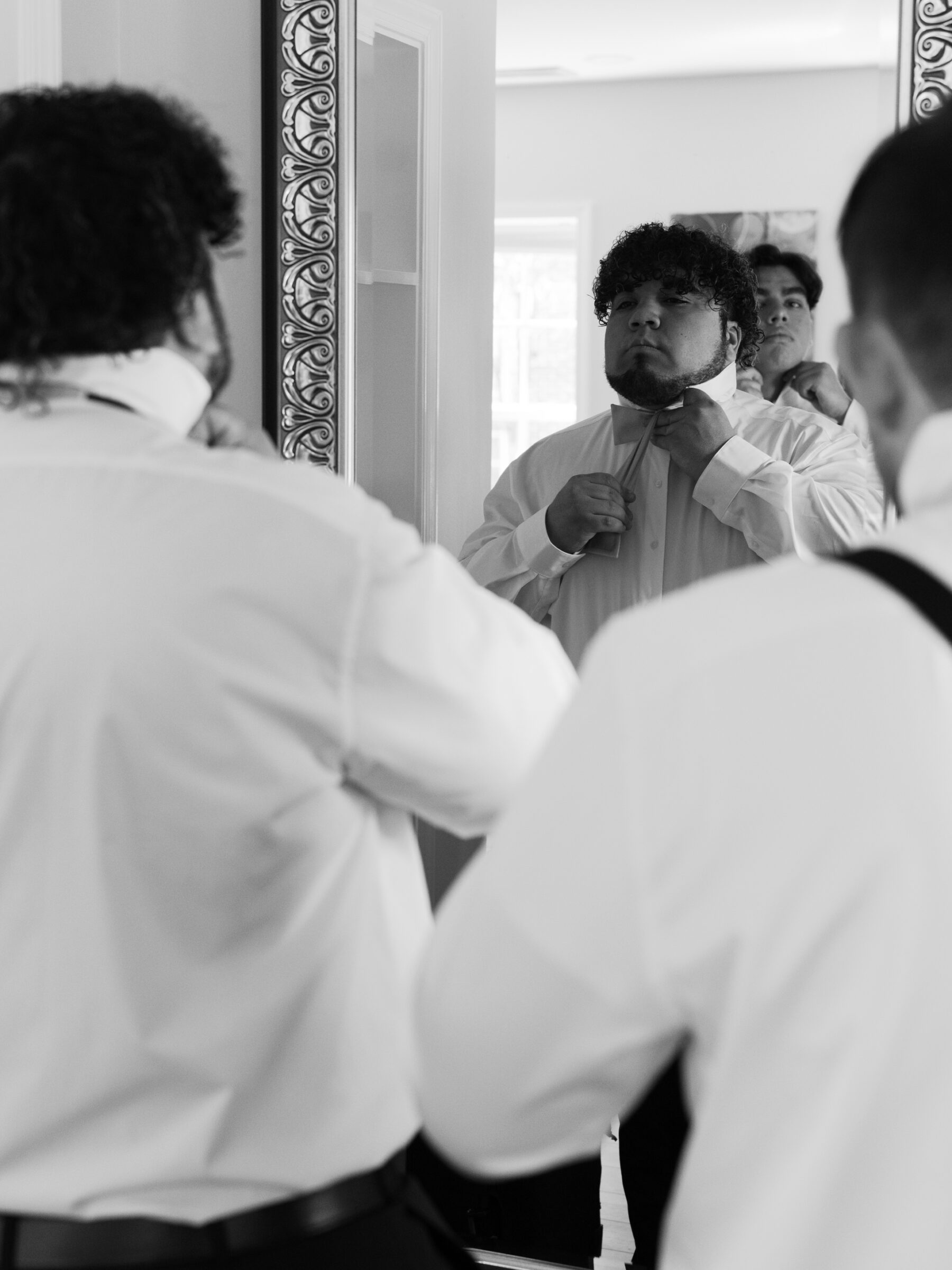 A black and white photo of a groom adjusting his bow tie in front of a mirror, while two groomsmen, dressed in white shirts and black suspenders, assist in the background. The reflection in the mirror captures the intimate, behind-the-scenes moment before the wedding ceremony.