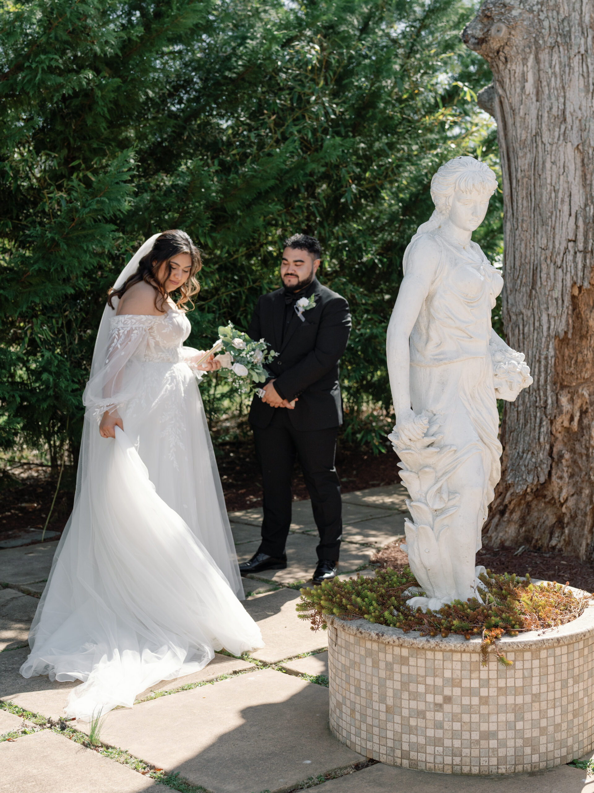 The bride, wearing an off-shoulder white wedding gown, gazes down as she adjusts her bouquet. The groom stands nearby, facing her with a gentle smile. In the foreground, a white marble statue of a woman holding flowers stands next to a flower bed, surrounded by greenery.
