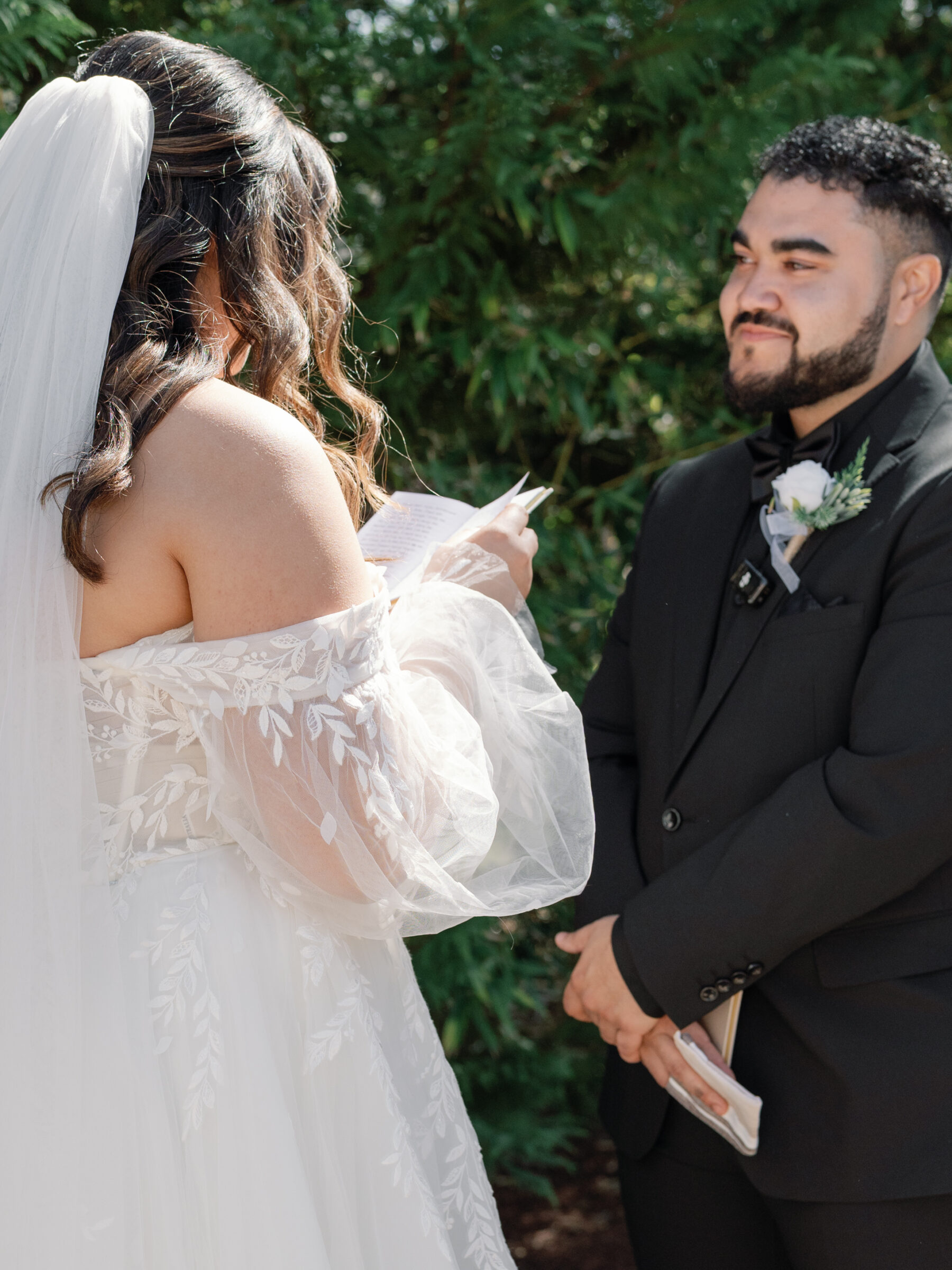 The bride, with her back to the camera, holds a piece of paper as she reads her vows. Her off-shoulder wedding gown features intricate floral embroidery. The groom, dressed in a black suit and tie, stands in front of her, smiling warmly. The background shows lush green foliage, creating a serene outdoor setting.