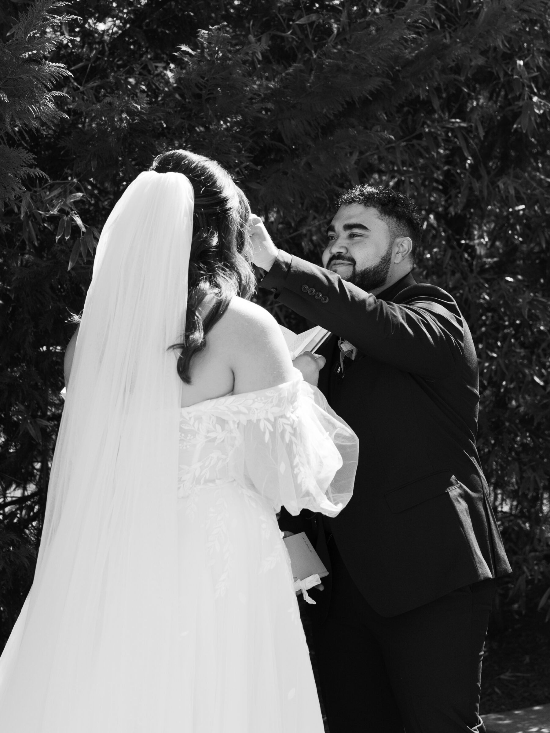 The groom gently wipes a tear from the bride’s cheek during their outdoor ceremony, surrounded by lush greenery. The bride, dressed in a soft, ethereal off-the-shoulder gown with delicate floral embroidery, gazes affectionately at her groom. The moment is captured in black and white, adding a timeless and emotional quality.