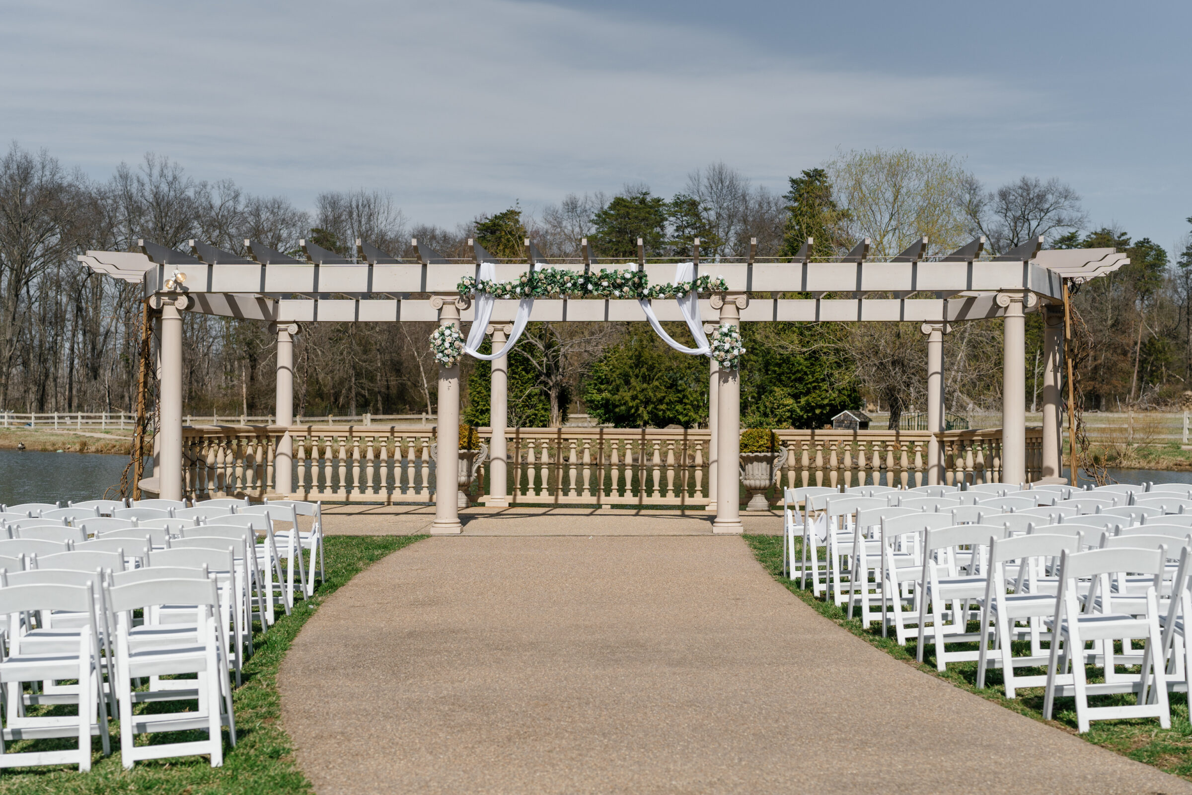 The outdoor wedding ceremony space at Morais Vineyards is set with rows of white folding chairs leading up to an elegant pergola adorned with floral garlands and flowing white fabric. The ceremony site is beautifully positioned by a tranquil water view, surrounded by lush trees and greenery, offering a serene and romantic atmosphere for the couple’s special day.