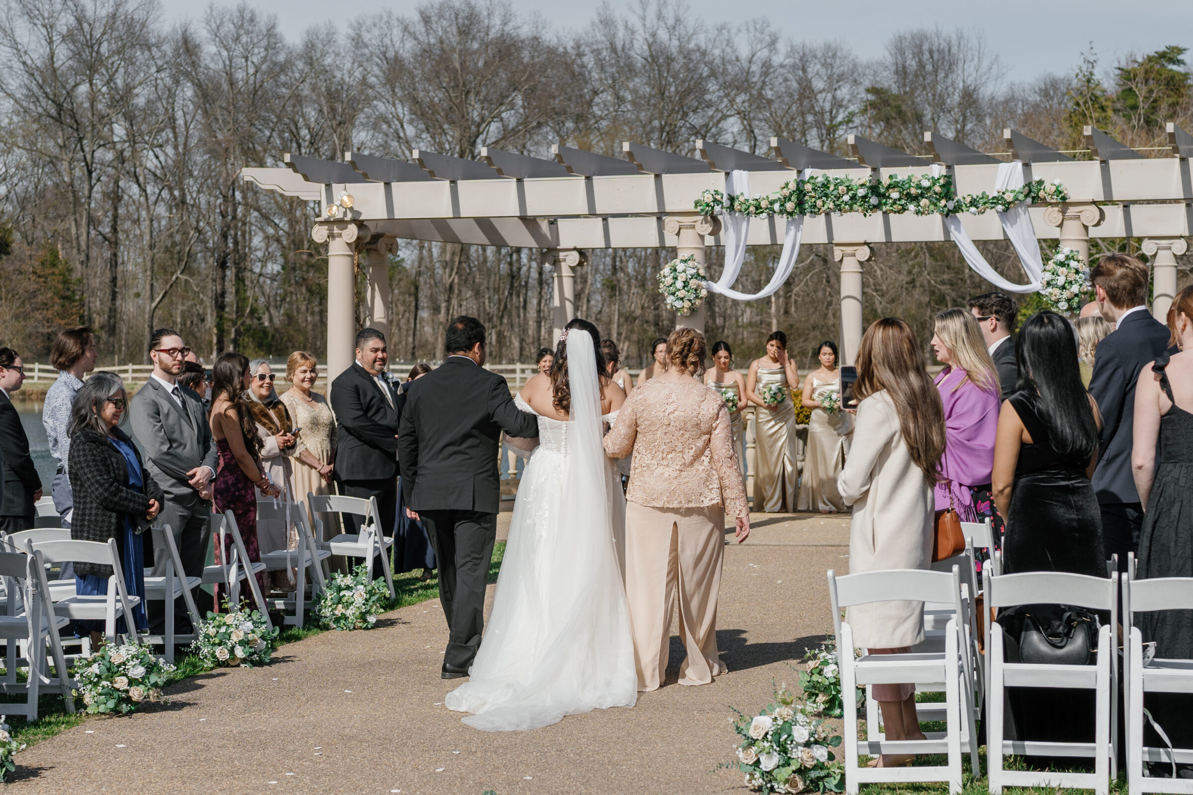 The bride, walking down the aisle with both parents, approaches the altar at Morais Vineyards. The groom and bridal party, including bridesmaids in gold dresses, await at the front, with the guests looking on. The floral decorations and the pergola above create a romantic and serene atmosphere.