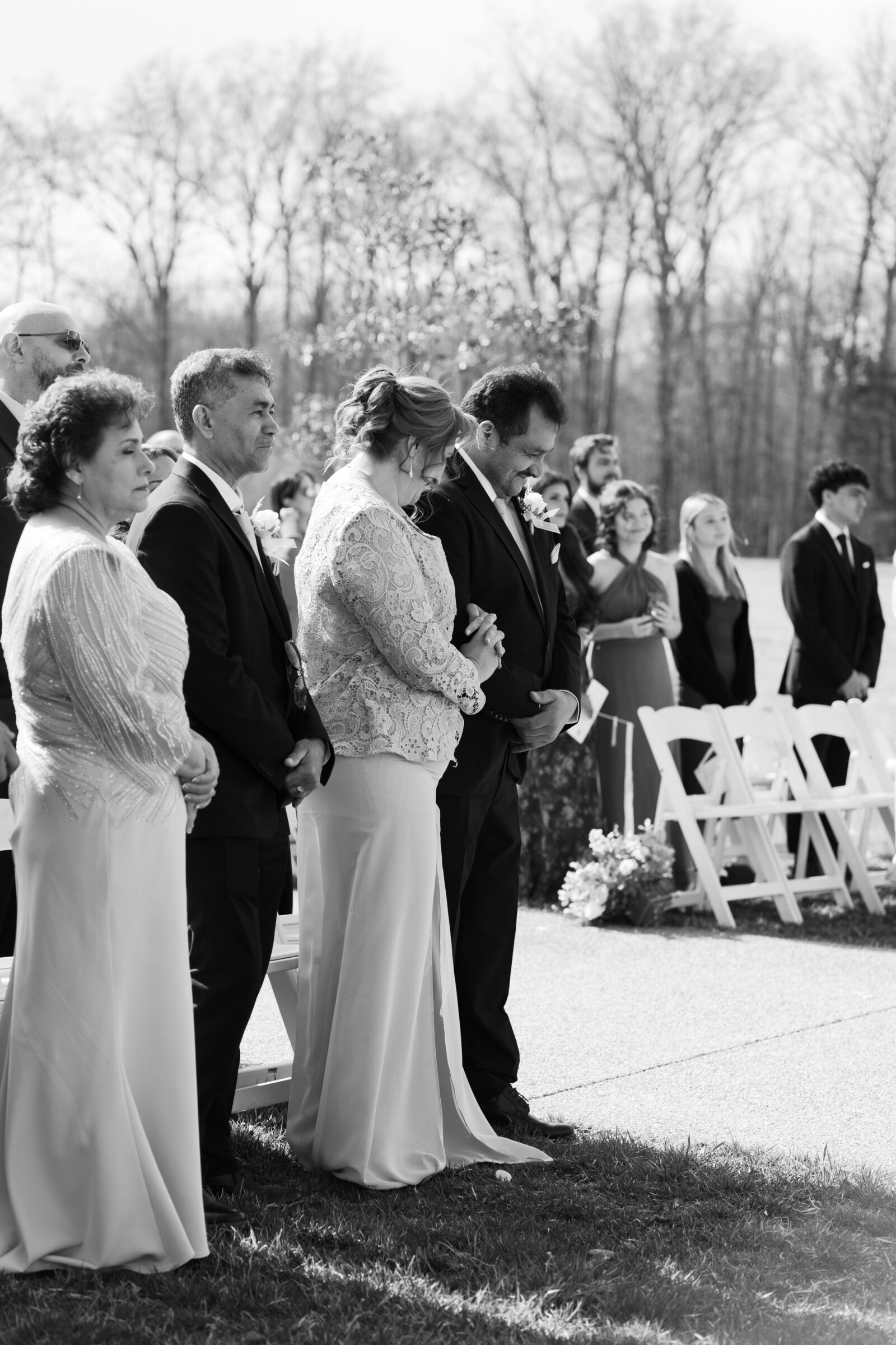 A black-and-white photo of wedding guests at the Morais Vineyards ceremony, including the bride’s parents and other family members, standing with their heads bowed and hands clasped in a moment of reflection. The soft sunlight and blurred background give the scene a peaceful, solemn feel.