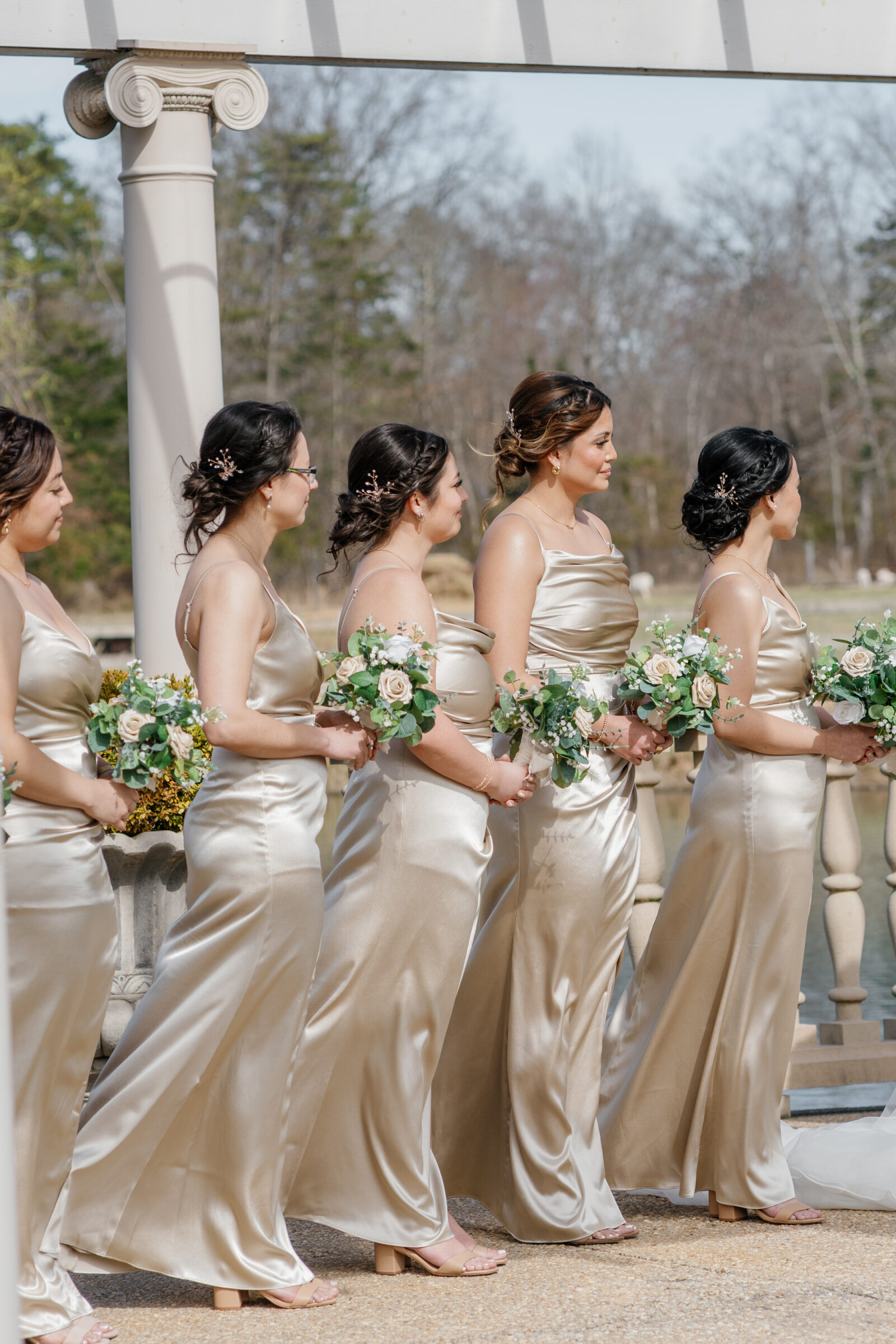 A line of bridesmaids in elegant satin champagne-colored gowns stands at the altar at Morais Vineyards, each holding a bouquet of white and blush roses. Their hair is styled in soft, romantic updos, with delicate hair accessories complementing their attire. The sun casts a soft glow on the serene outdoor setting.