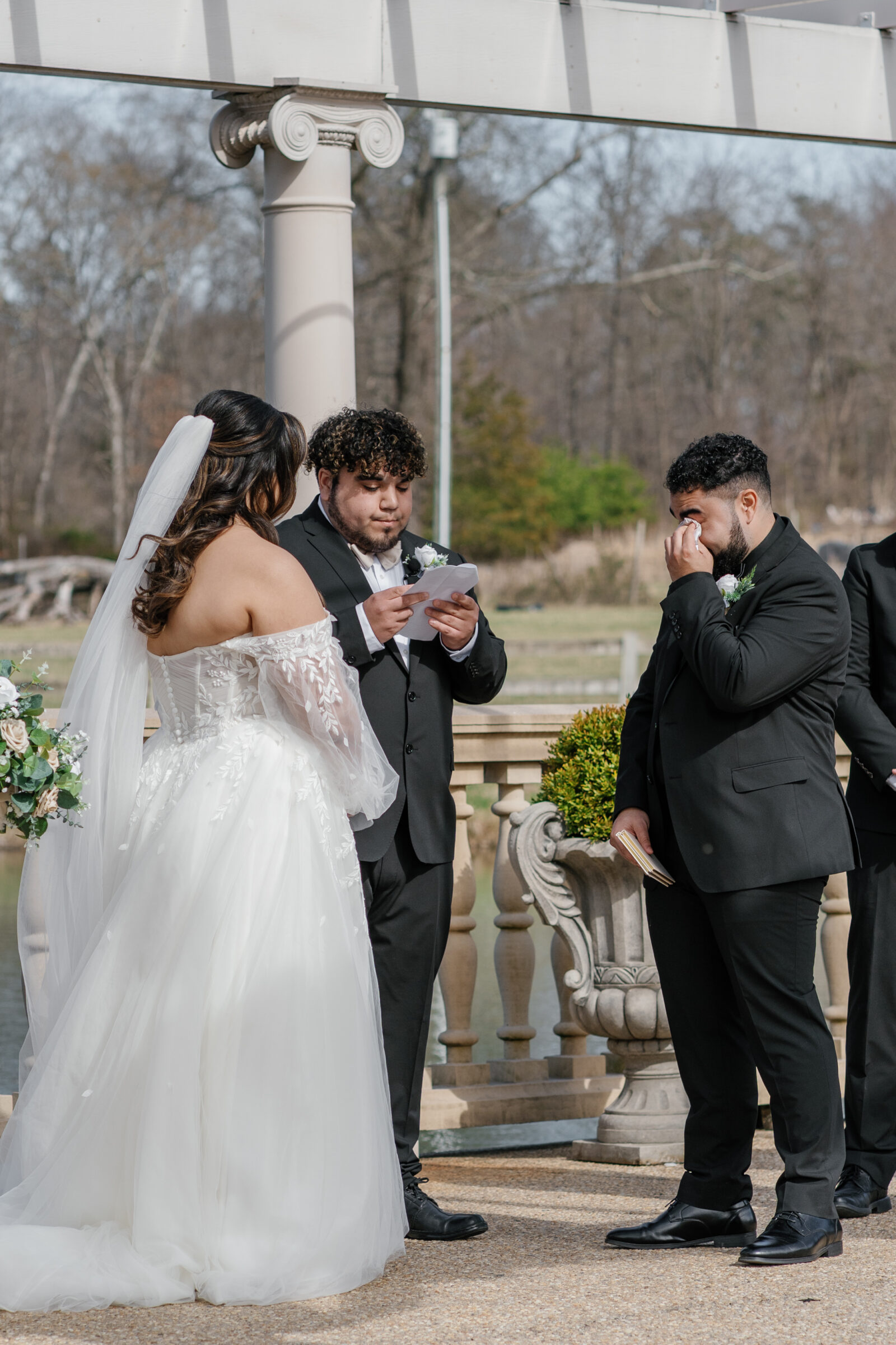 The groom, overcome with emotion, wipes away a tear while his best man reads from a note during the wedding ceremony at Morais Vineyards. The bride stands before him, holding a bouquet of white and blush roses, listening intently. The serene outdoor setting by the water adds to the intimate moment shared between the couple.