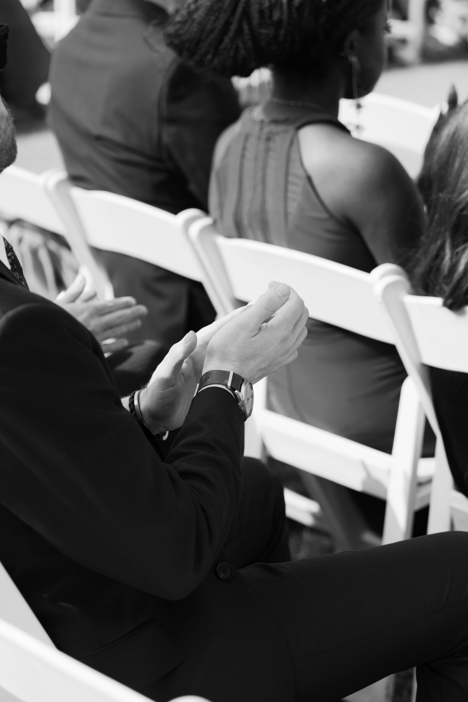 A close-up shot of a wedding guest's hands, with one hand clapping and the other resting on their lap. The guest wears a stylish watch and sits on a white folding chair, with the background featuring blurred figures of other attendees at the ceremony.