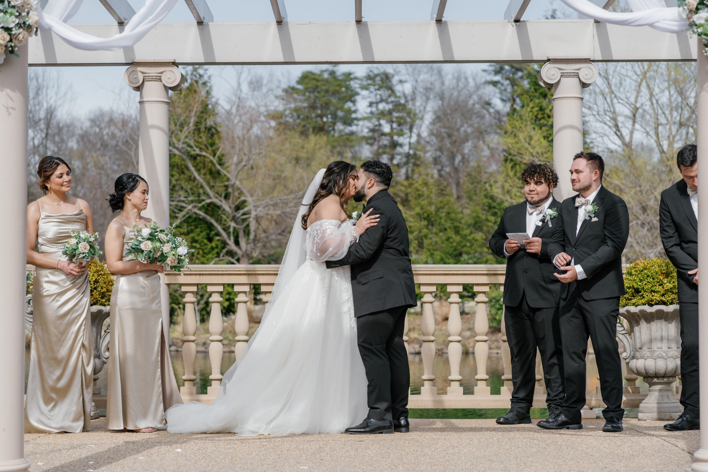 The bride and groom share their first kiss as newlyweds during their wedding ceremony at Morais Vineyards. The bride, dressed in a romantic off-the-shoulder gown, embraces her husband, who is wearing a classic black suit. In the background, bridesmaids in elegant satin dresses and groomsmen stand with smiles, witnessing the special moment beneath a beautifully decorated ceremony arch.