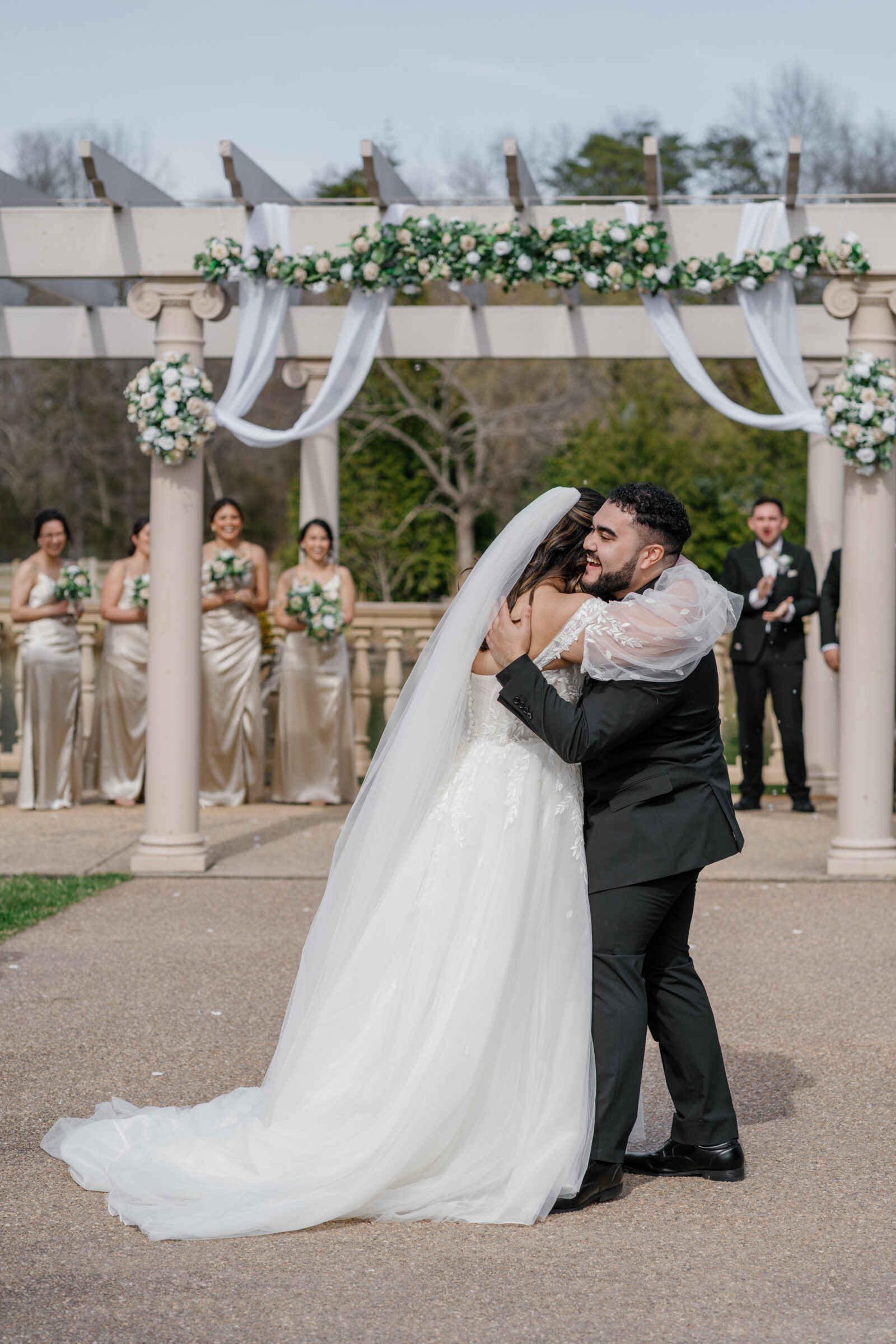 A newlywed couple shares a joyful hug during their wedding ceremony at Morais Vineyards. The bride in a flowing gown with delicate floral details embraces the groom, who is dressed in a classic black suit. In the background, the bridal party stands, smiling and celebrating the moment. The ceremony arch is decorated with lush greenery and white flowers.