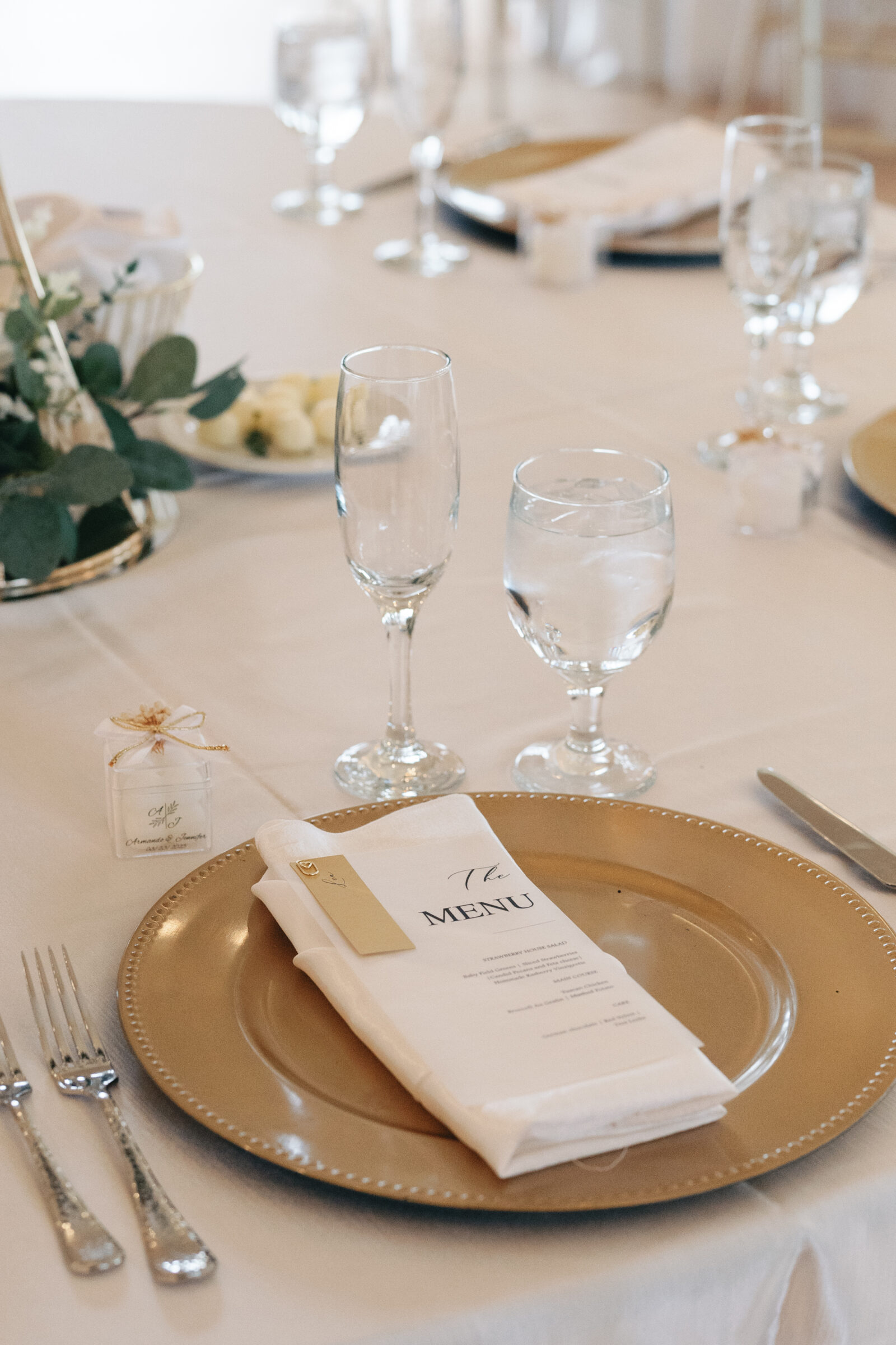 A close-up of a wedding reception table, featuring a gold charger plate, water glasses, and a small wedding favor box tied with ribbon. The menu is neatly folded on the napkin.