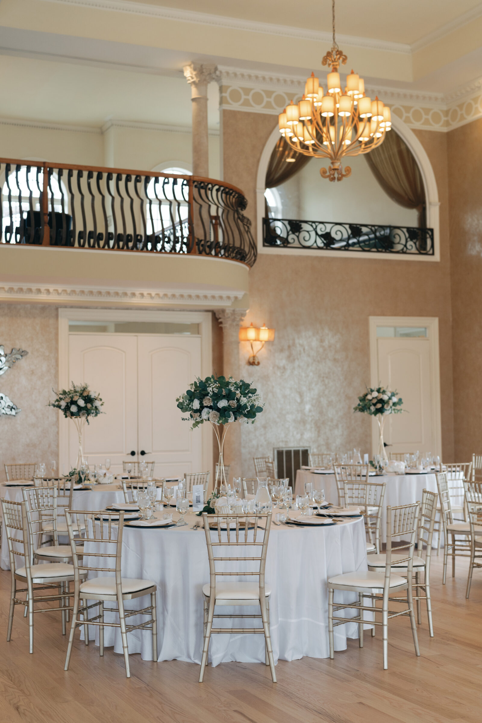 An interior shot of the grand wedding reception at Morais Vineyards, with multiple tables set for guests. Floral arrangements stand atop tall golden stands, and the chandelier overhead adds to the sophisticated atmosphere. The large, arched windows allow natural light to flood the room.