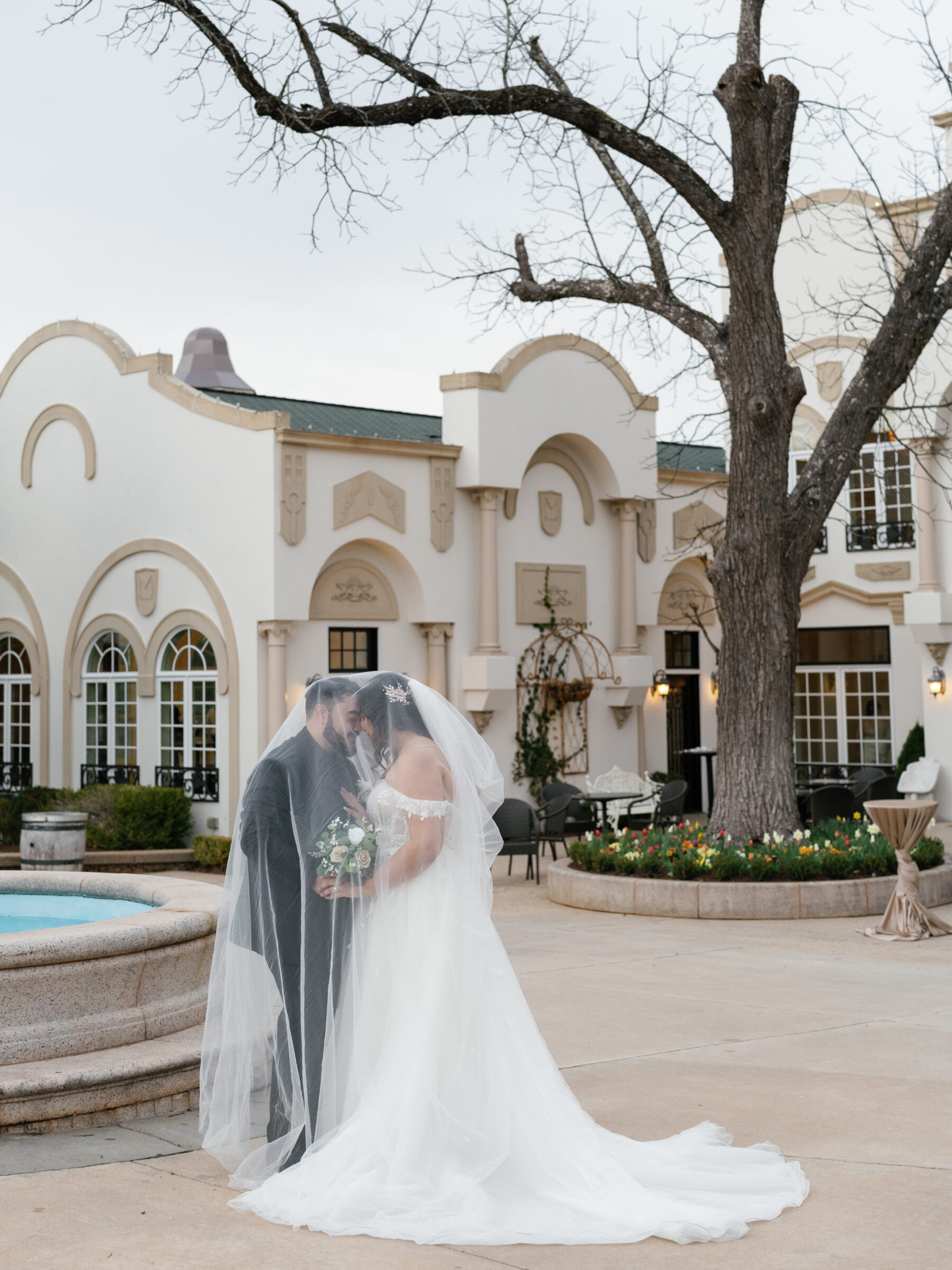 The bride and groom share a tender moment, their foreheads touching beneath the bride’s long veil, which cascades down around them. The groom holds the bride's bouquet gently while the background showcases a grand building and a bare tree. The soft colors and intimacy of the moment reflect a serene and romantic atmosphere.