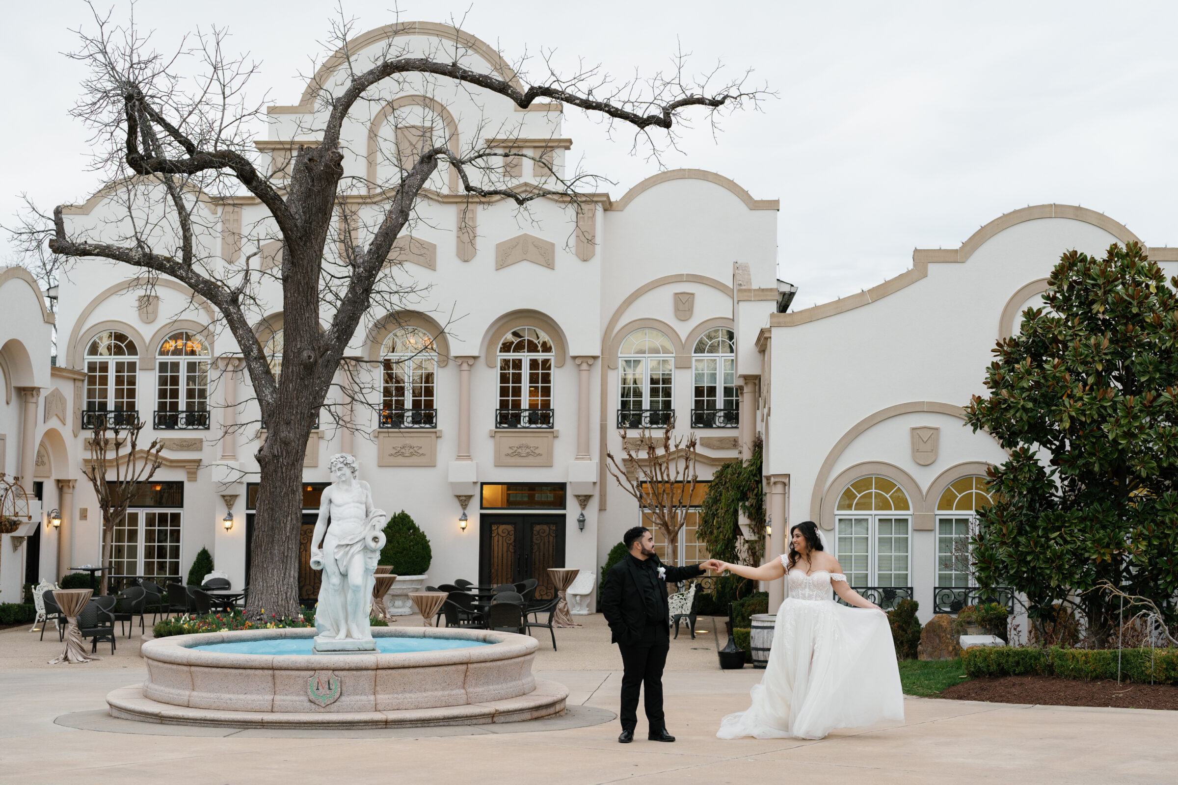 The bride and groom share a joyful moment outside Morais Vineyards, with the bride playfully twirling her dress while the groom smiles at her. Behind them stands a classical fountain and a bare tree, complementing the elegant architecture of the venue. The light and airy scene captures their carefree happiness.