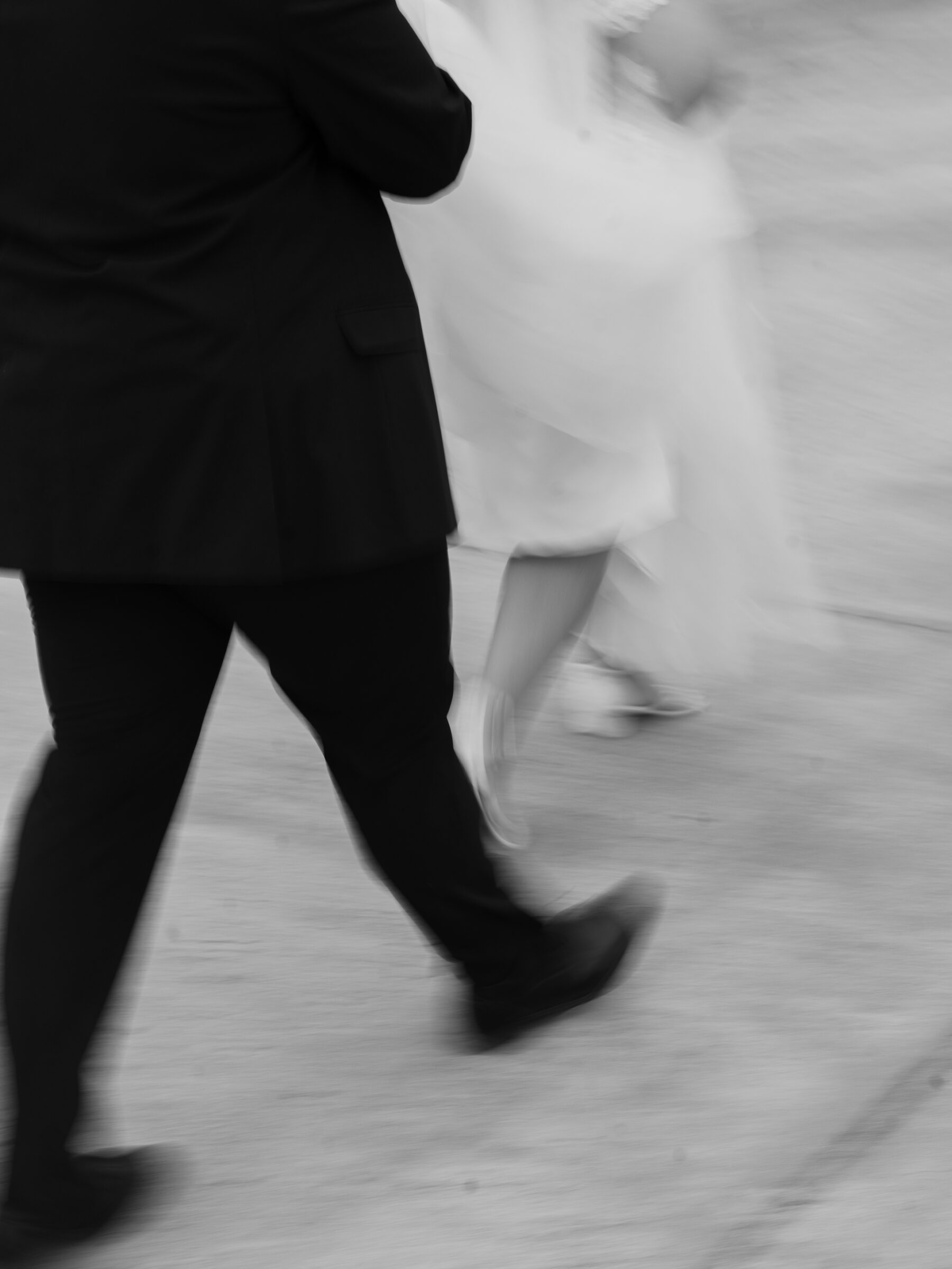 A blurred, black-and-white photograph capturing the bride and groom walking together. The groom, dressed in a black suit, is seen holding the bride's hand while she walks beside him, her wedding dress flowing behind her. The motion blur adds a sense of movement and excitement, emphasizing the couple’s intimate moment as they stroll together.