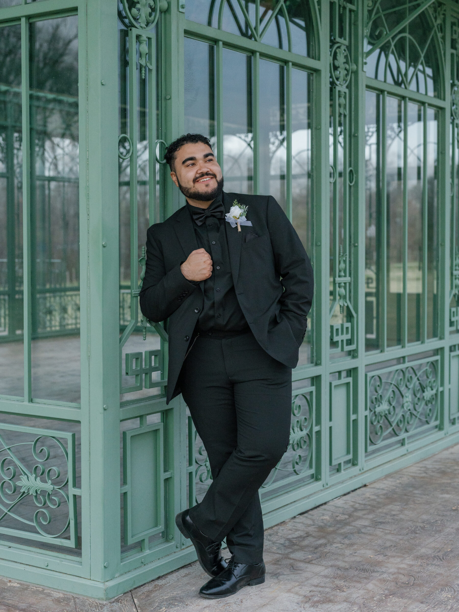Groom standing confidently in front of the green conservatory at Morais Vineyards, Bealeton, Virginia. He is dressed in a classic black tuxedo, leaning casually against the window frame with a smile.