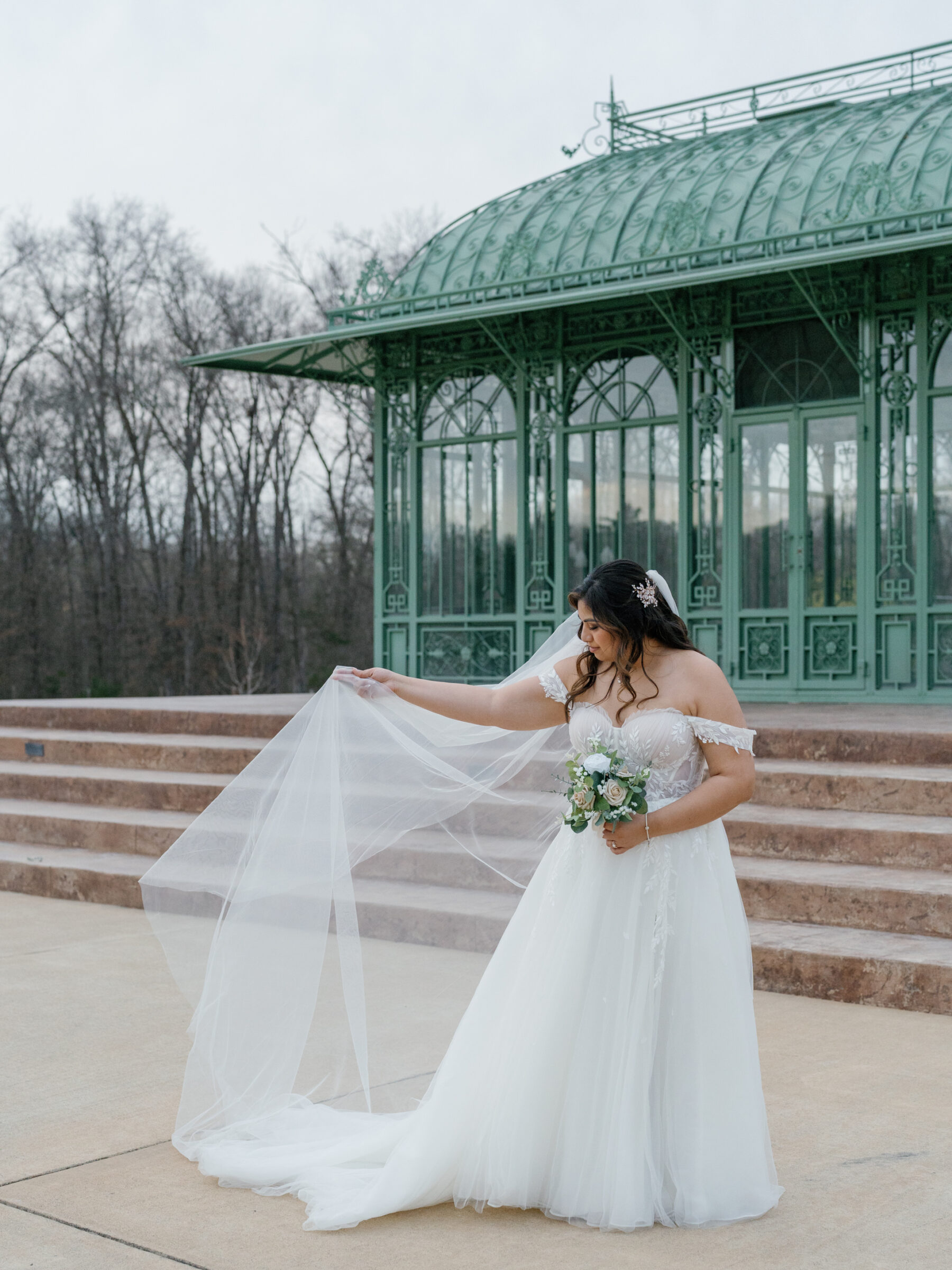 Bride adjusting her veil, standing in front of the green conservatory at Morais Vineyards, Bealeton, Virginia. Her flowing gown and delicate bouquet of white and blush roses complete the elegant look.