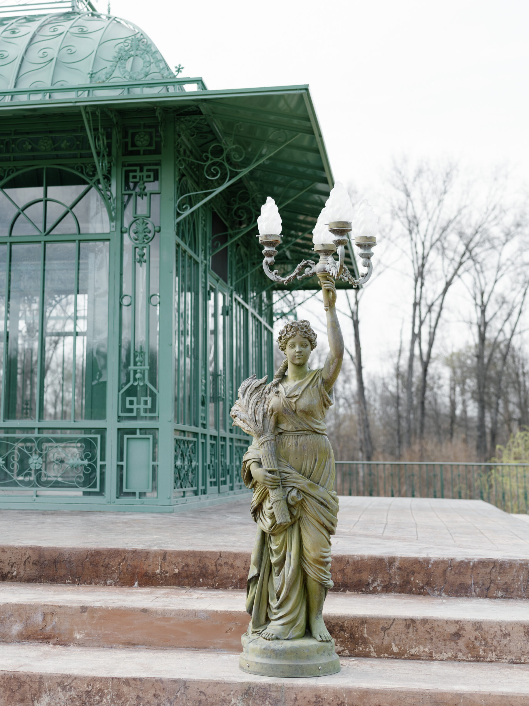 A decorative statue holding a candelabra stands near the gazebo at Morais Vineyards, surrounded by a rustic stone platform and winter trees in the background.