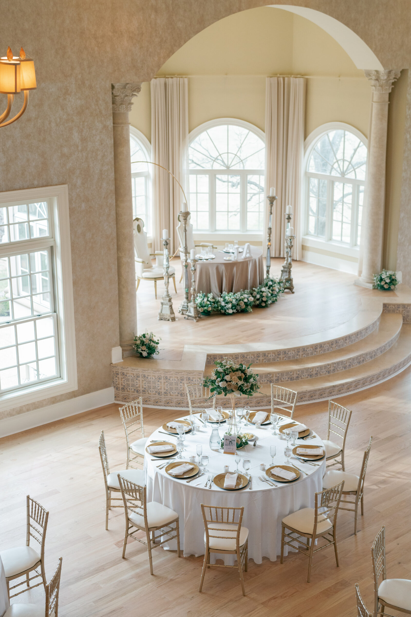 An elegant reception space at Morais Vineyards with a round table set for guests in the foreground and the bride and groom's table decorated with flowers in the background, framed by grand windows.
