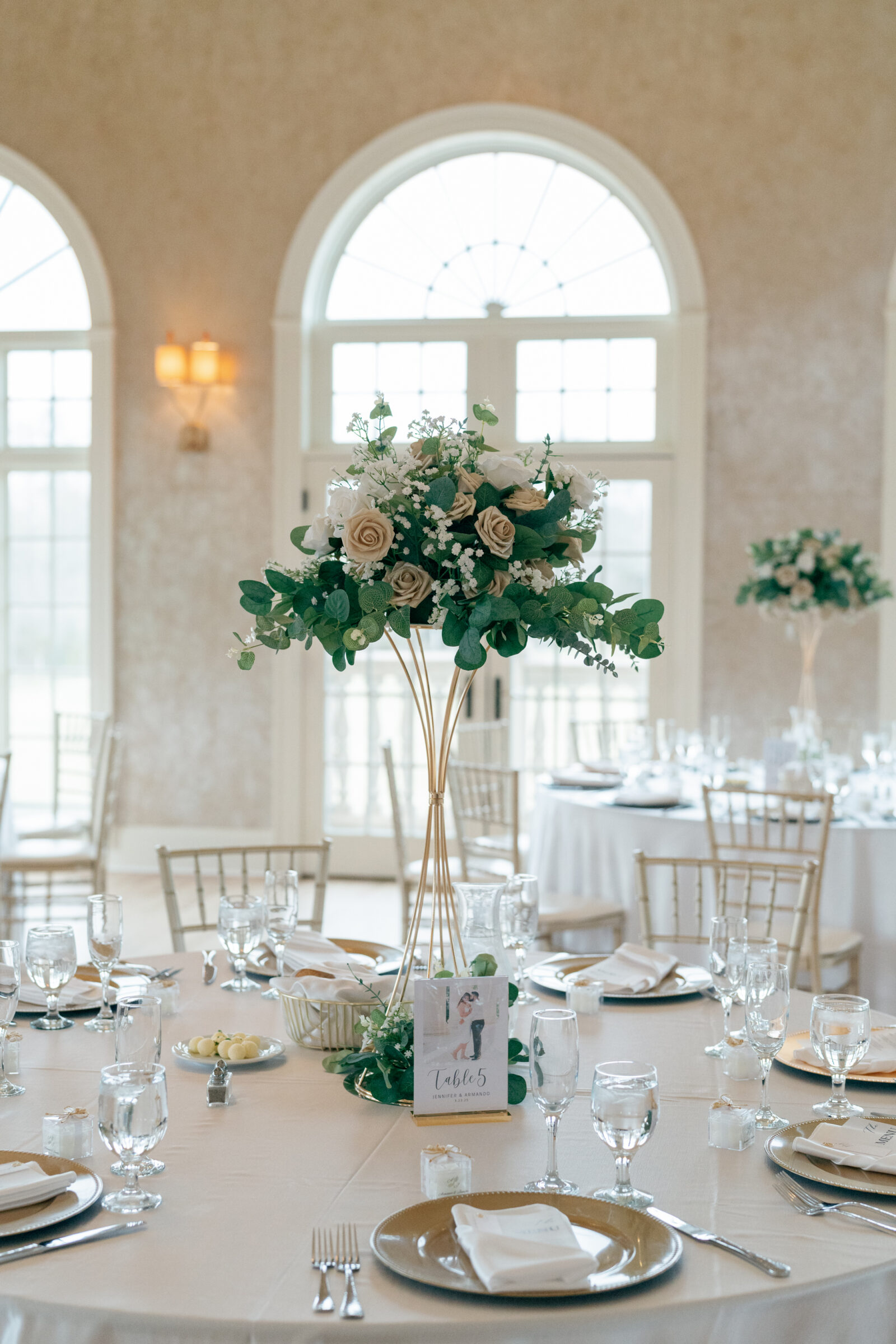 Elegant wedding reception table at Morais Vineyards featuring a tall floral centerpiece with white and blush roses, greenery, and a gold base. The table is set with glassware, plates, and silverware, with a table number card and small guest favors.