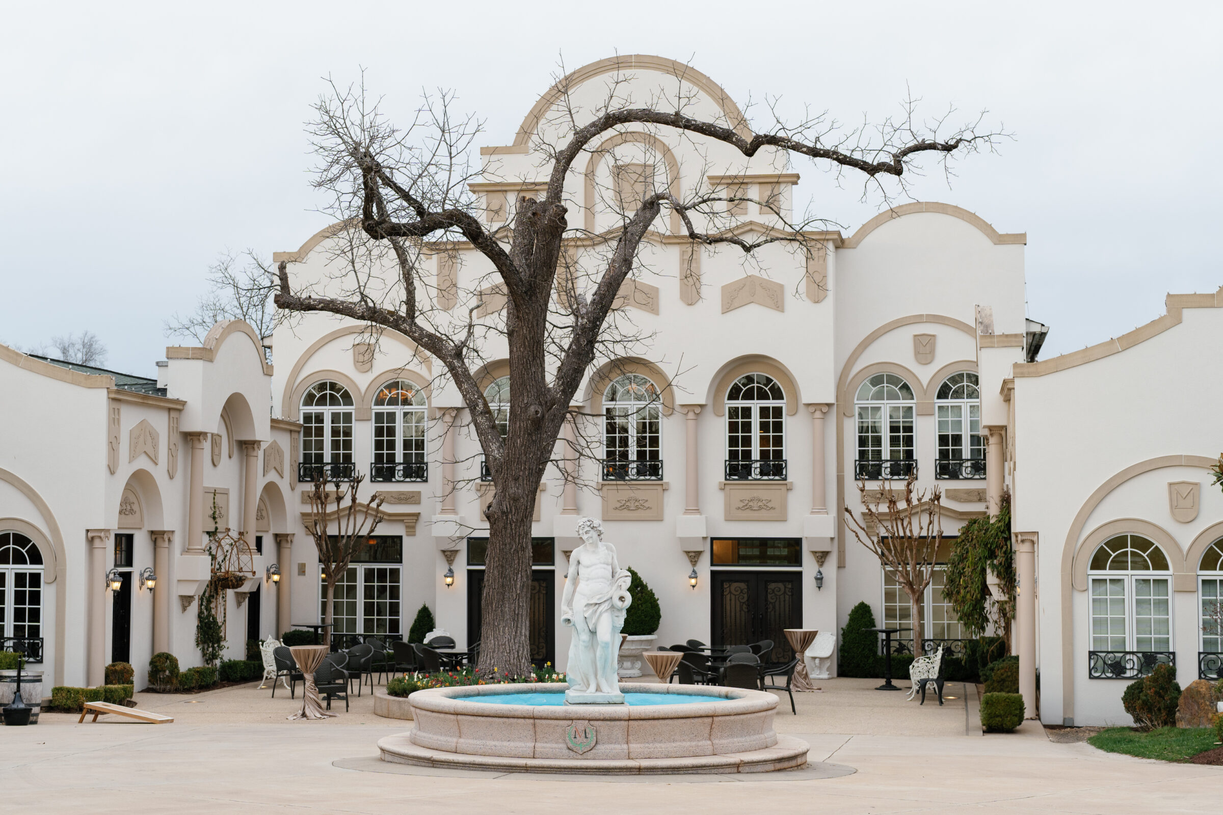 Exterior view of Morais Vineyards in Bealeton, VA, featuring a beautiful fountain with a statue in the center, surrounded by manicured trees and the venue's grand white architecture. The courtyard is adorned with outdoor seating, creating an elegant atmosphere.