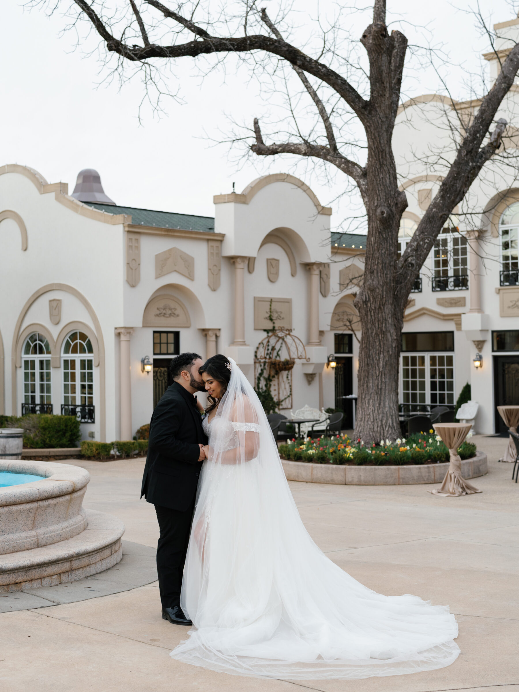 A romantic couple stands by the fountain in the courtyard of Morais Vineyards in Bealeton, VA, sharing a quiet, intimate moment. The bride, wearing a flowing wedding gown with a long veil, embraces her groom, with the vineyard's elegant architecture and manicured landscaping in the background.