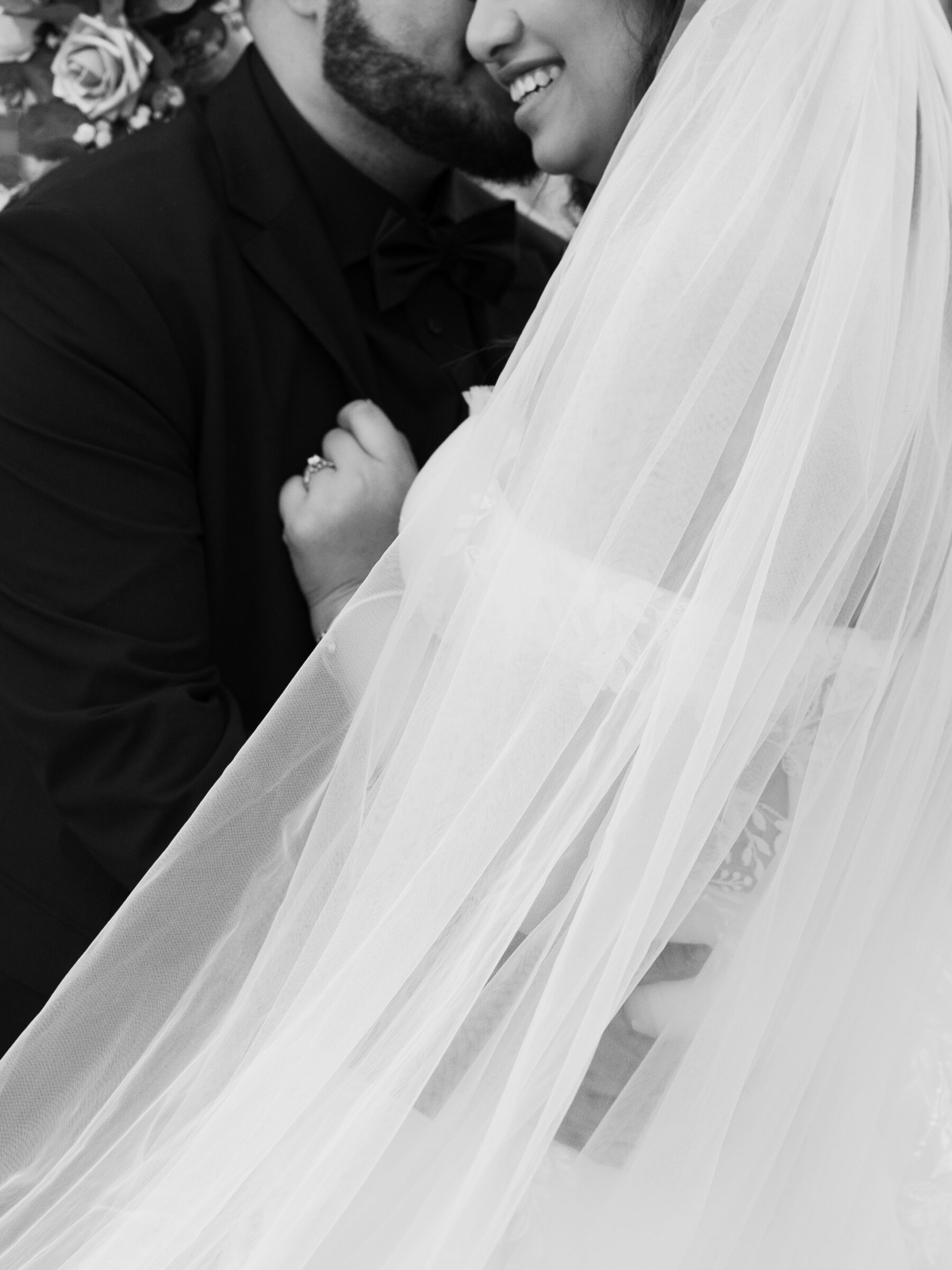 A close-up, black and white image of a couple sharing an intimate moment at their wedding. The bride, in her delicate wedding veil, smiles as she gently holds her groom’s suit, while the groom is seen with a soft expression, his face near hers. The background features romantic, blurred floral details.