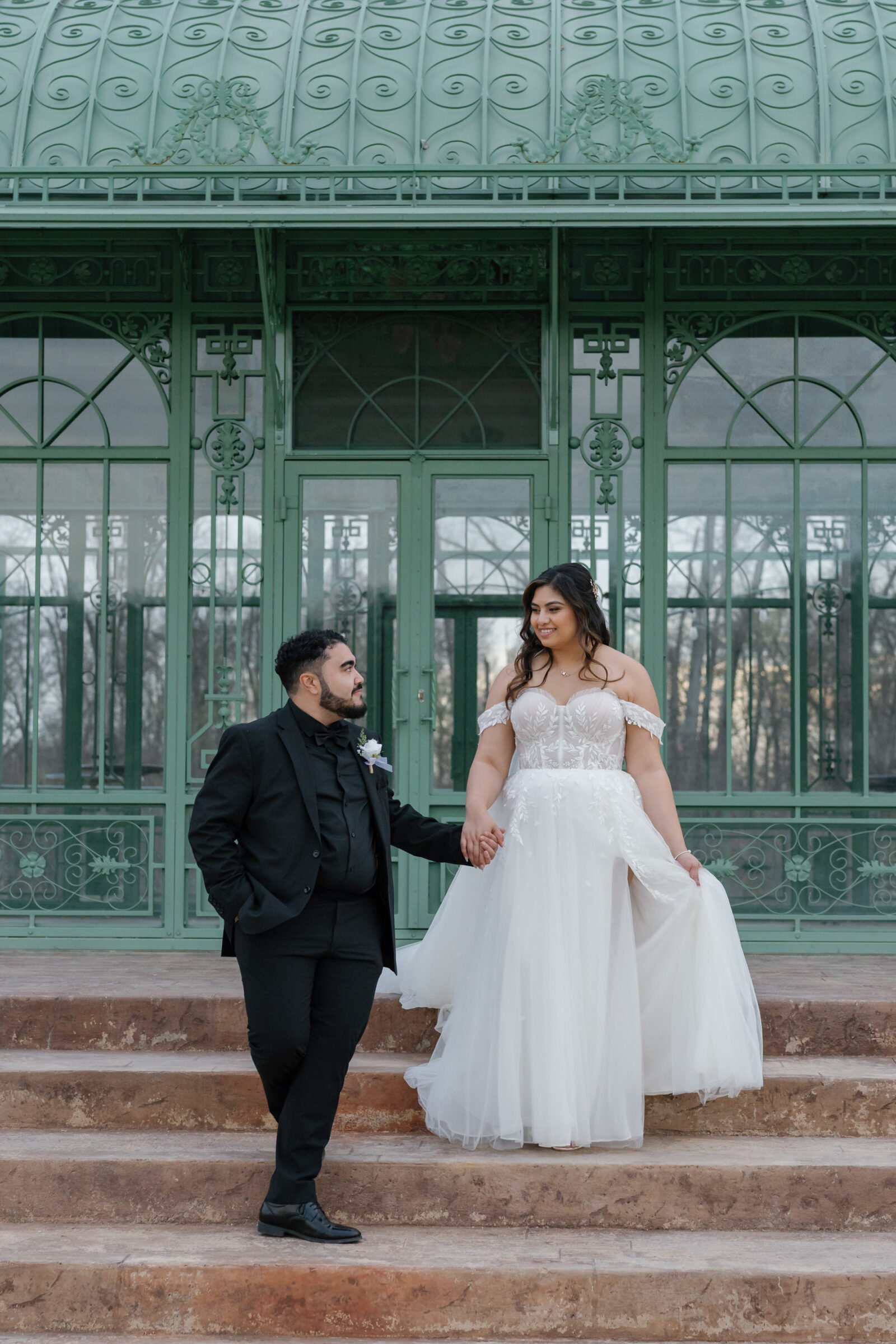 A joyful newlywed couple standing hand in hand on the steps of a beautiful green gazebo at Morais Vineyards. The bride, wearing a flowing off-the-shoulder wedding gown, smiles at her groom, who is dressed in a black tuxedo. The lush greenery and the intricate ironwork of the gazebo create an elegant, romantic backdrop for this intimate moment.