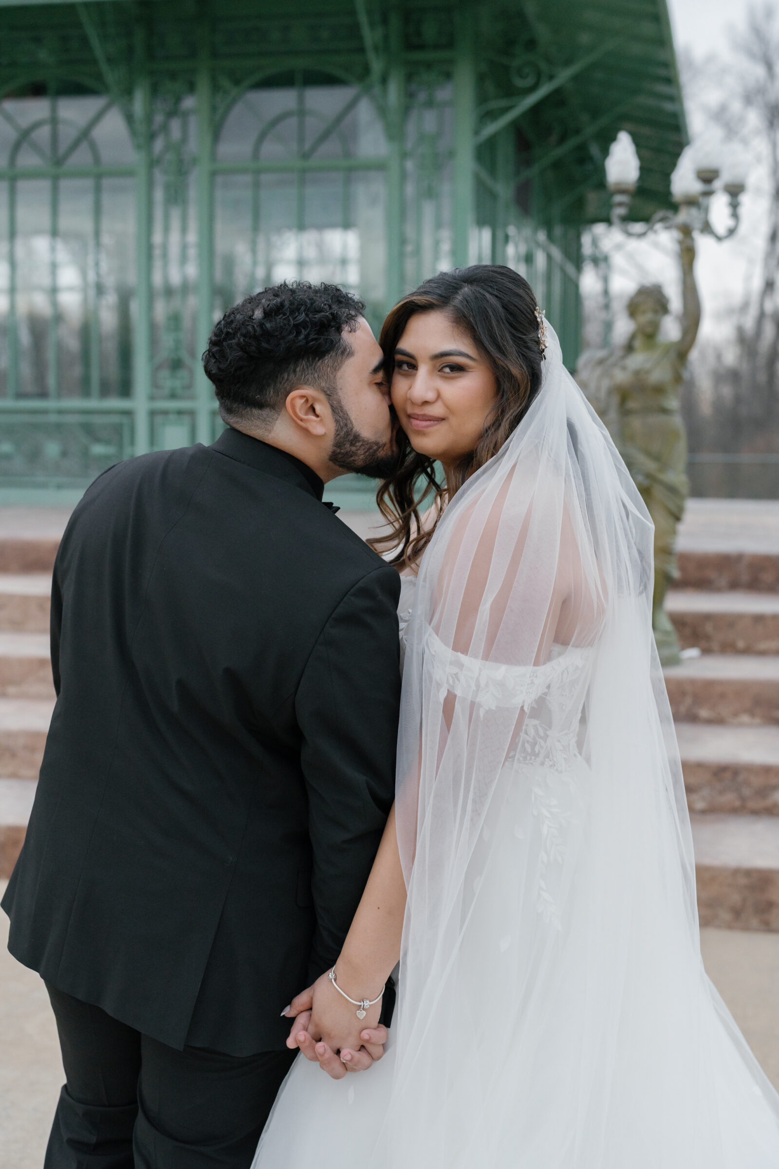 A loving moment captured as the groom kisses his bride's cheek while they stand together in front of a green iron gazebo at Morais Vineyards. The bride, in an off-the-shoulder wedding gown and veil, gazes at the camera with a gentle smile, while the groom stands beside her, holding her hand. A statue of a woman holding a light post stands in the background, enhancing the romantic ambiance.