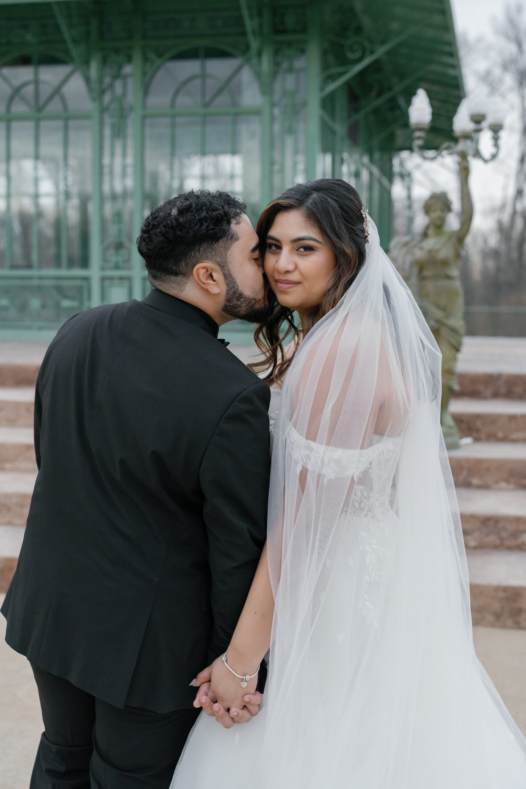 A loving moment captured as the groom kisses his bride's cheek while they stand together in front of a green iron gazebo at Morais Vineyards. The bride, in an off-the-shoulder wedding gown and veil, gazes at the camera with a gentle smile, while the groom stands beside her, holding her hand. A statue of a woman holding a light post stands in the background, enhancing the romantic ambiance.