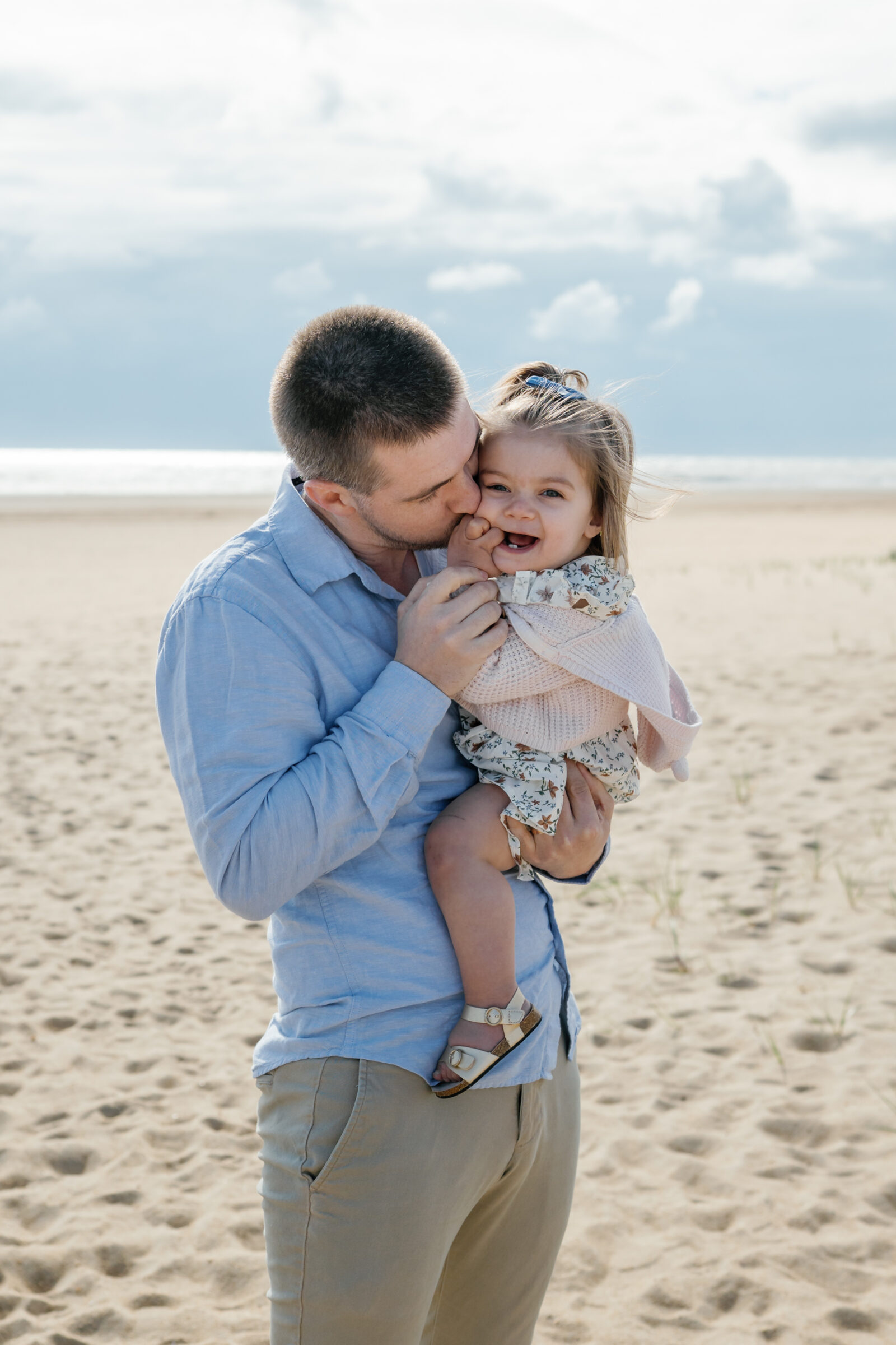 A father kissing his daughter on the cheek while holding her on the beach at North End Beach, Virginia Beach. The daughter is smiling with joy, her hand near her mouth.