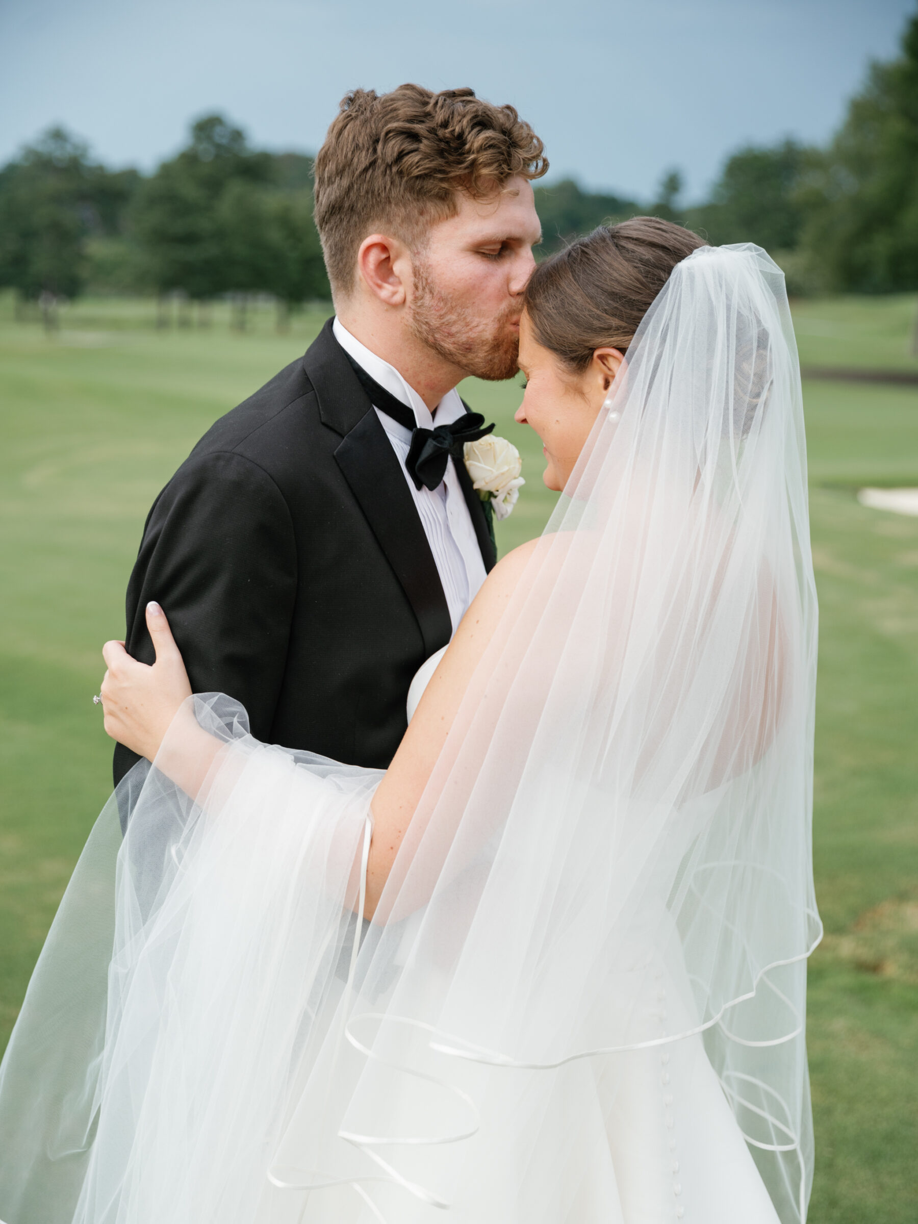 The groom lovingly kisses the bride's forehead, their heads gently touching as she smiles. The soft veil cascades over her shoulders, flowing down to her dress. The serene golf course stretches out behind them, adding to the intimate, quiet moment between the newlyweds.