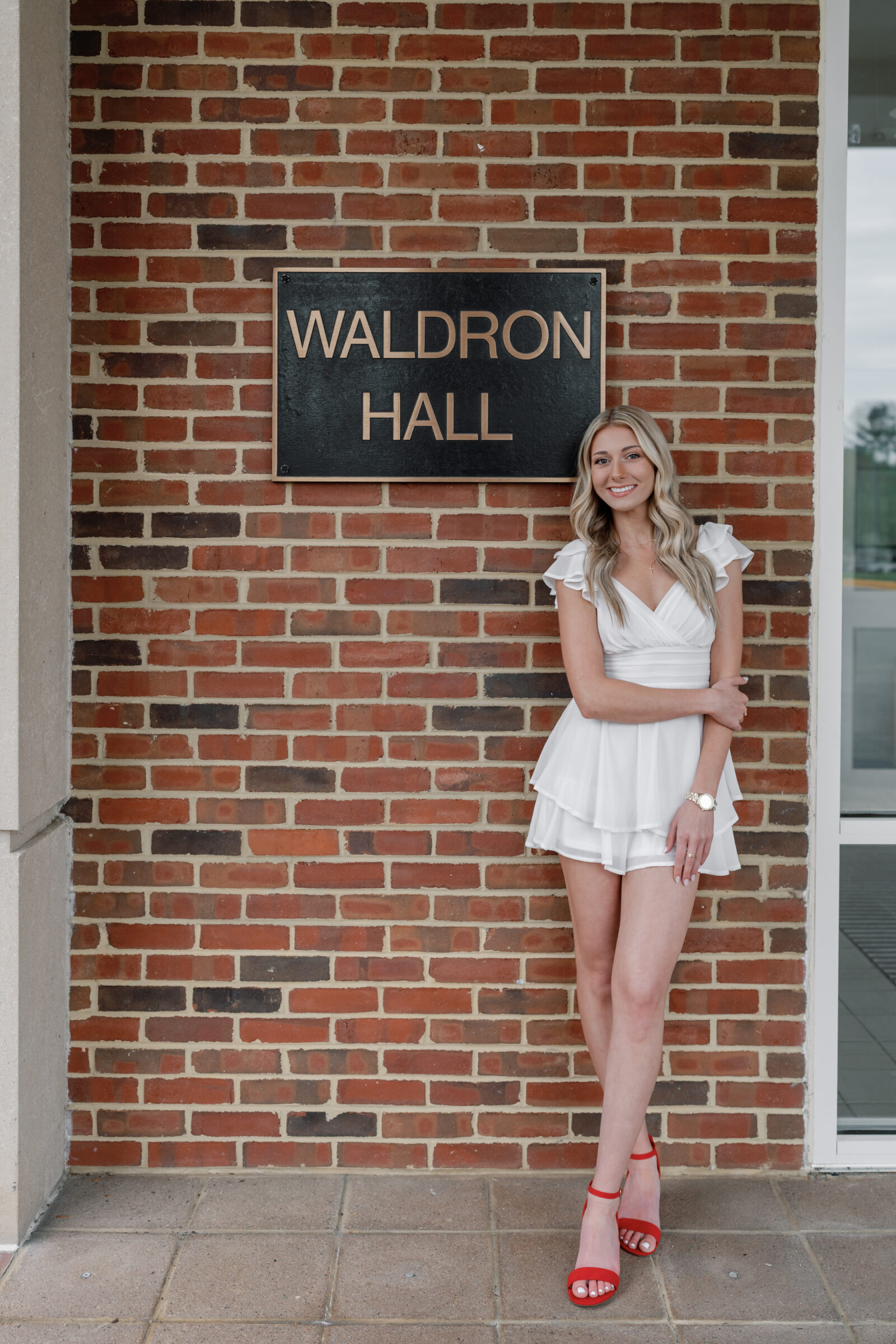 A smiling female senior standing in front of a brick wall with a "WALDRON HALL" sign at Radford University. She is wearing a white dress with a tiered skirt and red high heels, posing confidently with one arm gently resting on her waist. She has long, blonde hair and is accessorized with a watch and simple jewelry.
