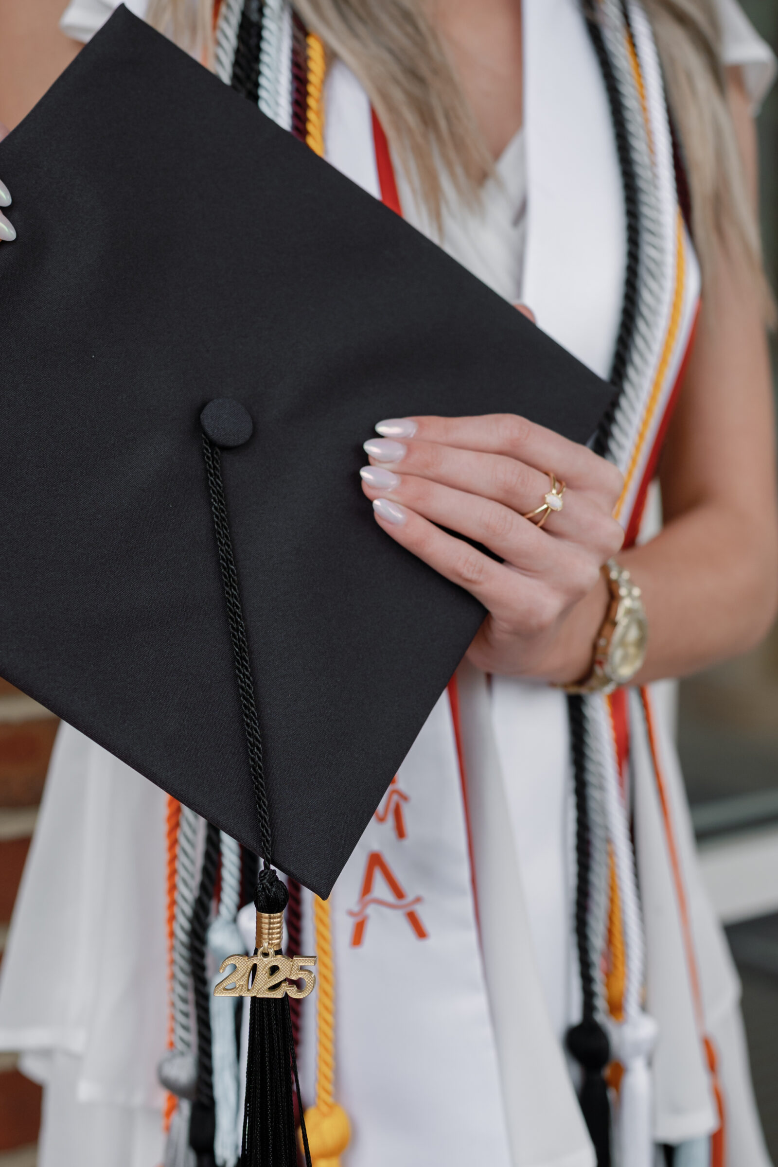 Close-up of a senior student holding a graduation cap with a gold '2025' tassel charm, showcasing colorful graduation cords and a delicate gold ring on their hand. The background features a brick wall, adding a personal and celebratory touch to the moment.