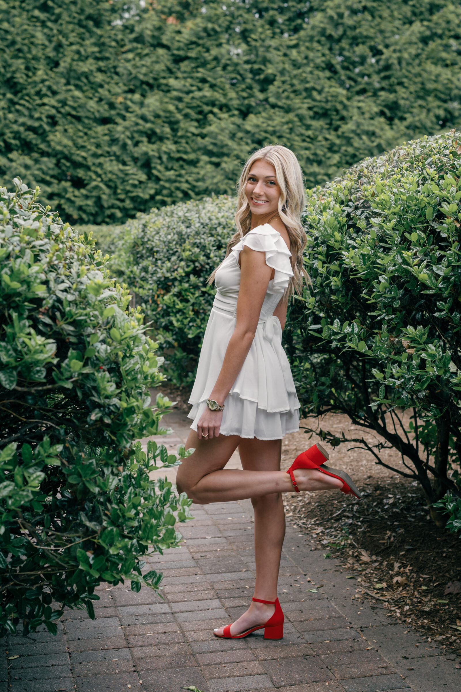 A senior student in a white ruffled dress and red heels, striking a playful pose in a lush garden at Radford University. She stands on one leg, smiling at the camera with her foot lifted, surrounded by green bushes and a tranquil path.