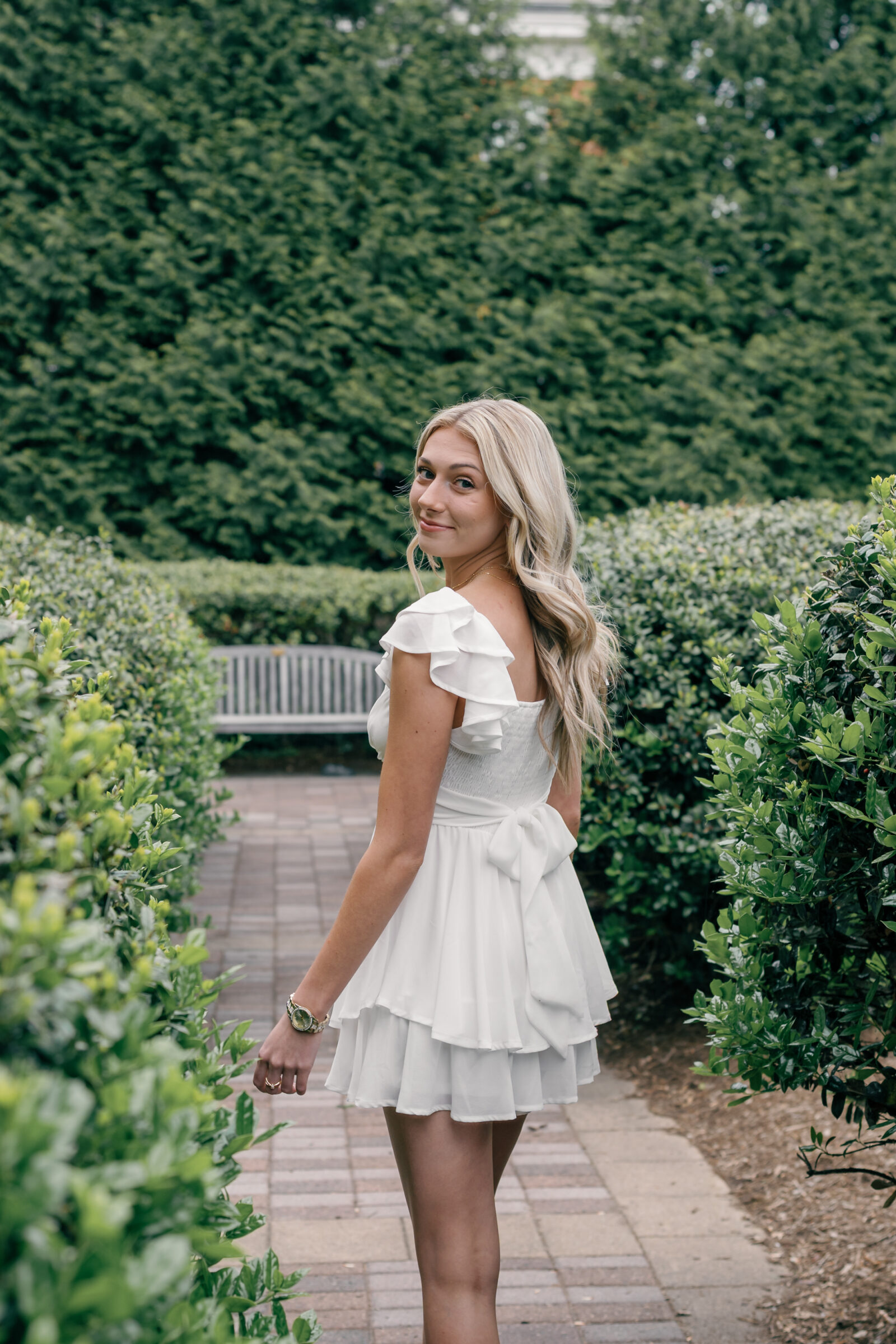 A senior student in a white ruffled dress, walking through a lush garden path at Radford University. She smiles over her shoulder, with soft waves in her blonde hair, a relaxed and joyful expression, and a glimpse of a bench in the background.