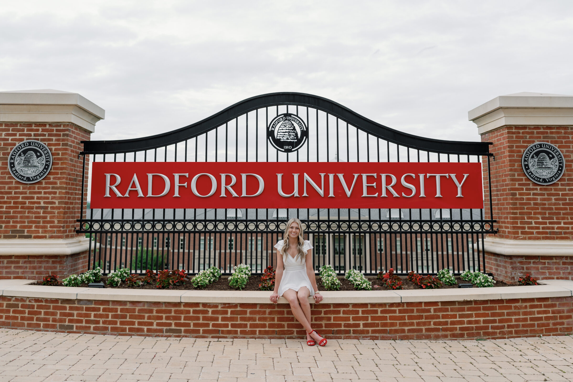 A senior student sitting on a brick wall in front of the iconic Radford University sign, wearing a white dress and red heels. She smiles at the camera, surrounded by colorful flowers and a cloudy sky.