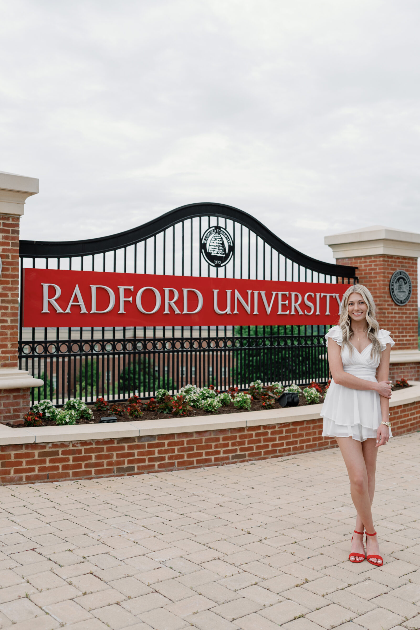 A senior student standing in front of the Radford University entrance sign, smiling and wearing a white ruffled dress and red heels. The scene includes colorful flowers and a cloudy sky in the background, highlighting the university’s iconic gate.