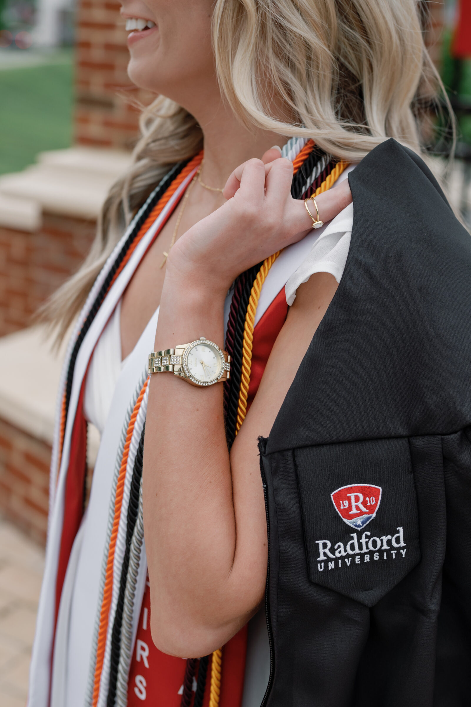 A senior student with blonde hair, wearing a gold watch and a graduation ring, casually drapes her Radford University cap and gown over her shoulder. She shows off her colorful graduation cords in a relaxed pose, with the Radford University emblem visible on the gown.