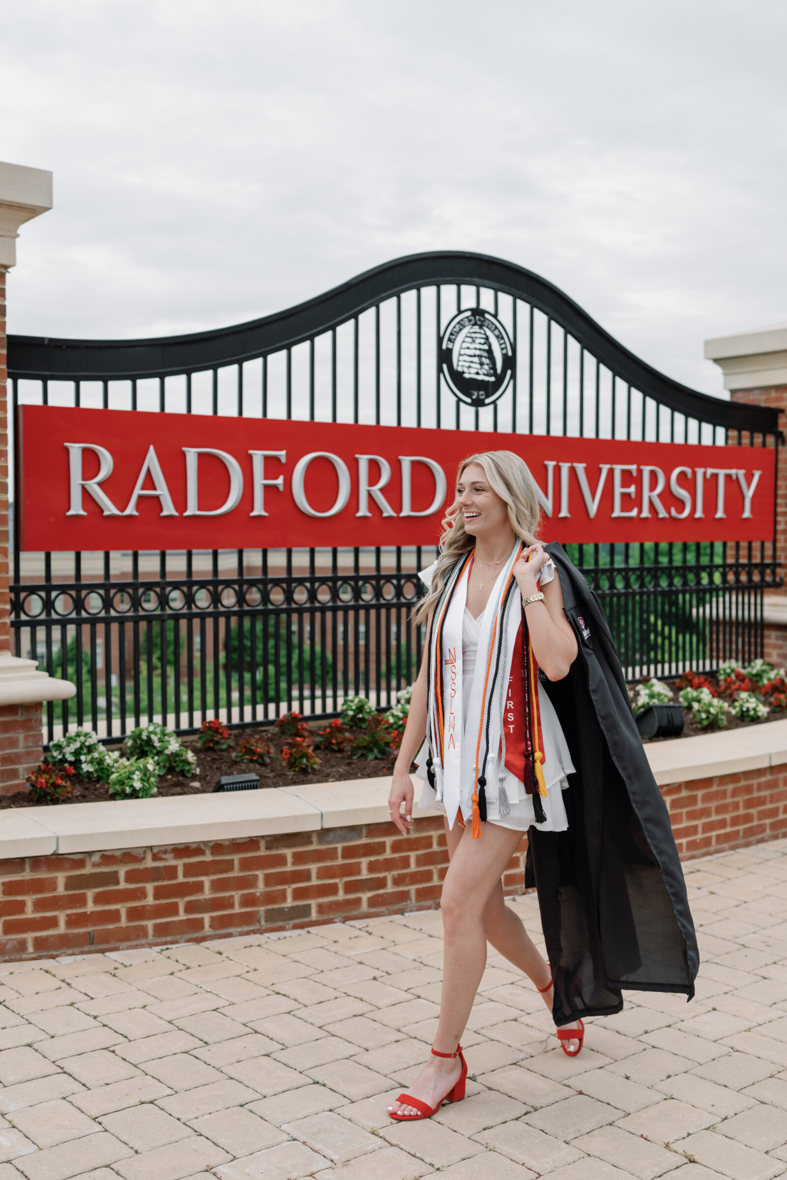 A senior student walking proudly in front of the Radford University entrance sign, wearing a white ruffled dress, red heels, and multiple graduation cords. She carries her cap and gown over her shoulder, smiling and enjoying the moment with flowers and the iconic university gate in the background.