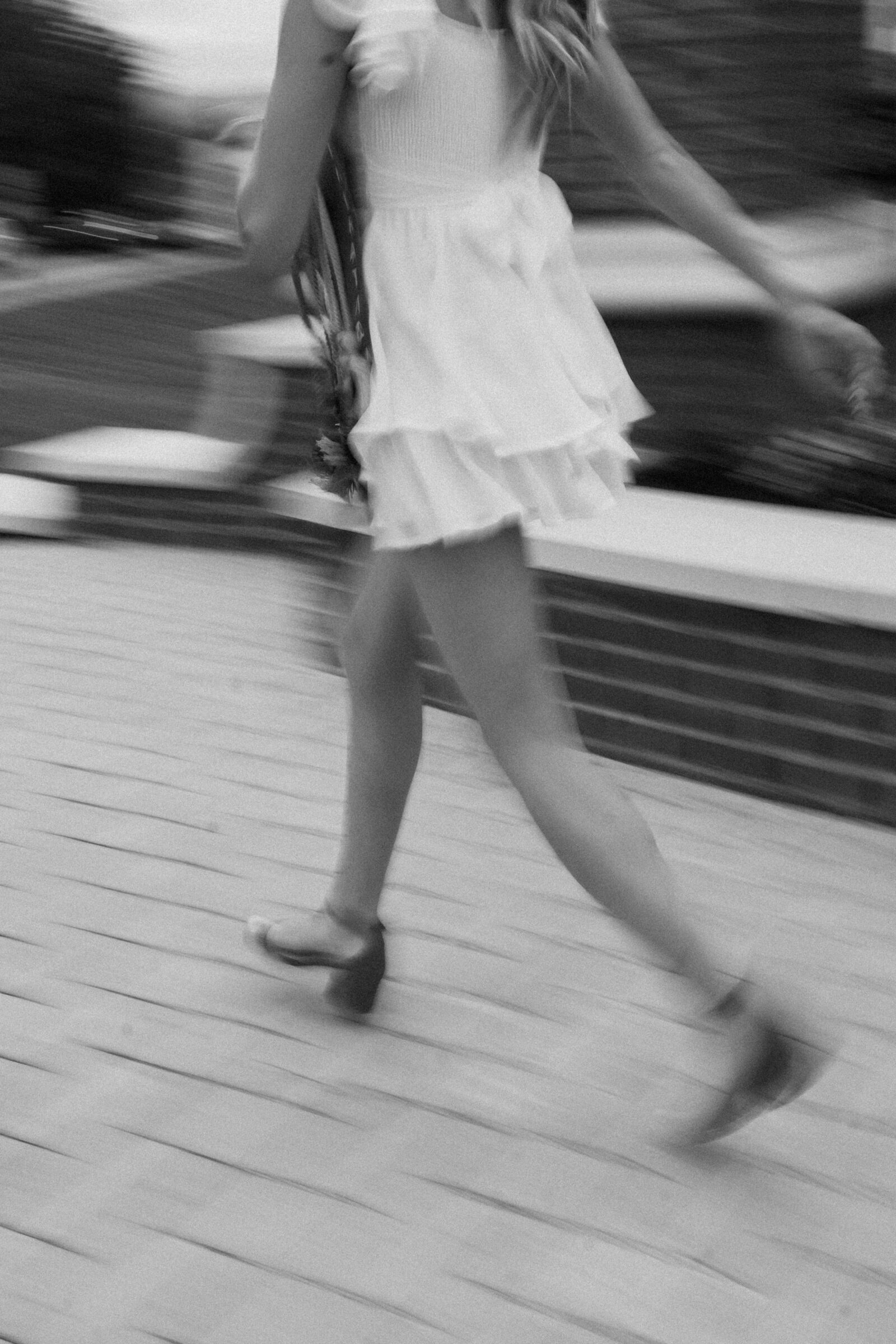 A motion blur of a senior student walking, wearing a white ruffled dress and red heels. The image captures a dynamic, candid moment as she moves through the Radford University campus, with soft movement in her legs and dress.