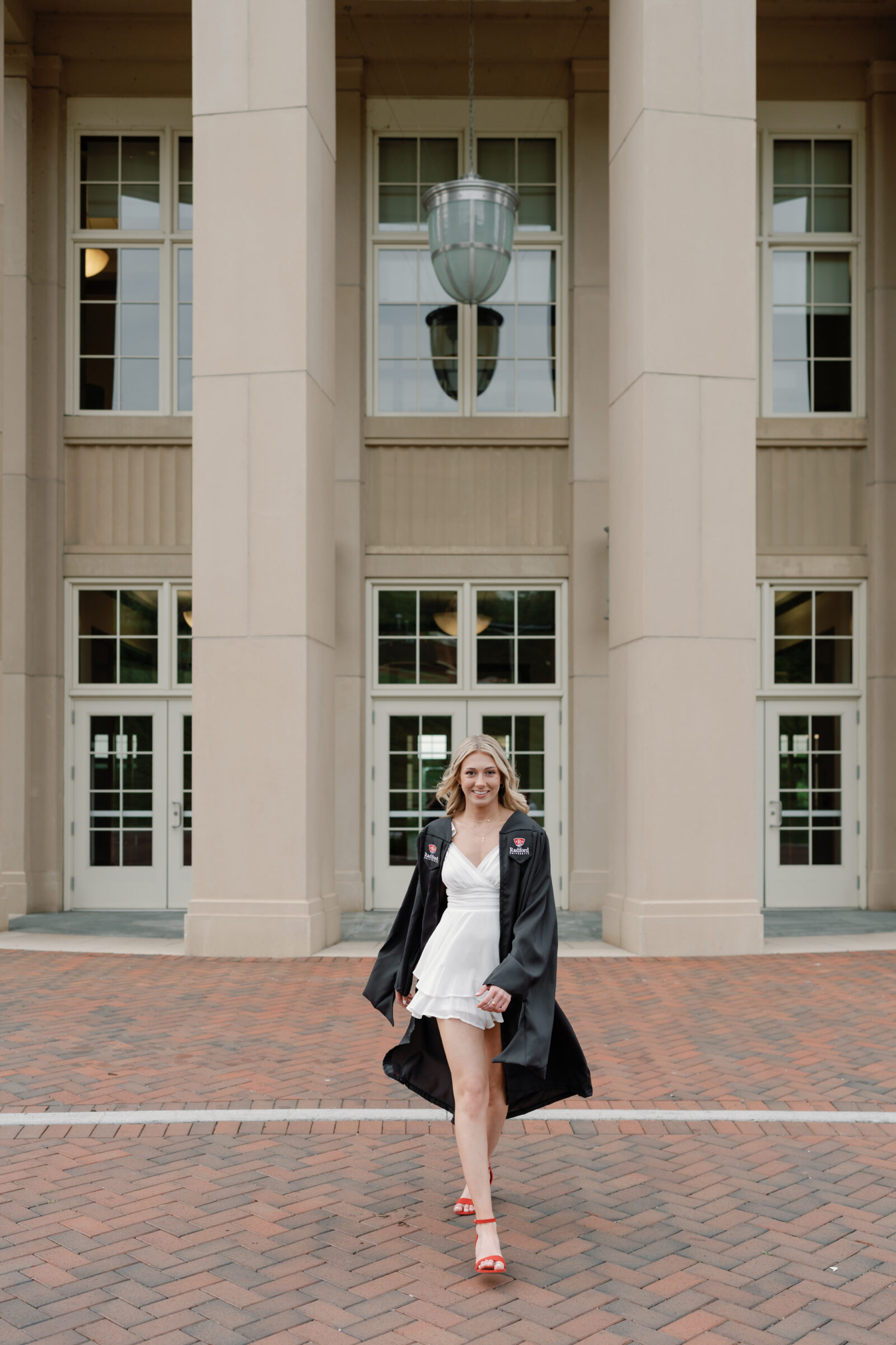 A senior student walking confidently in front of a large, classic university building. She wears a white ruffled dress and red heels, with her Radford University cap and gown draped over her shoulders. The image captures her joyful expression as she moves across the brick pathway.