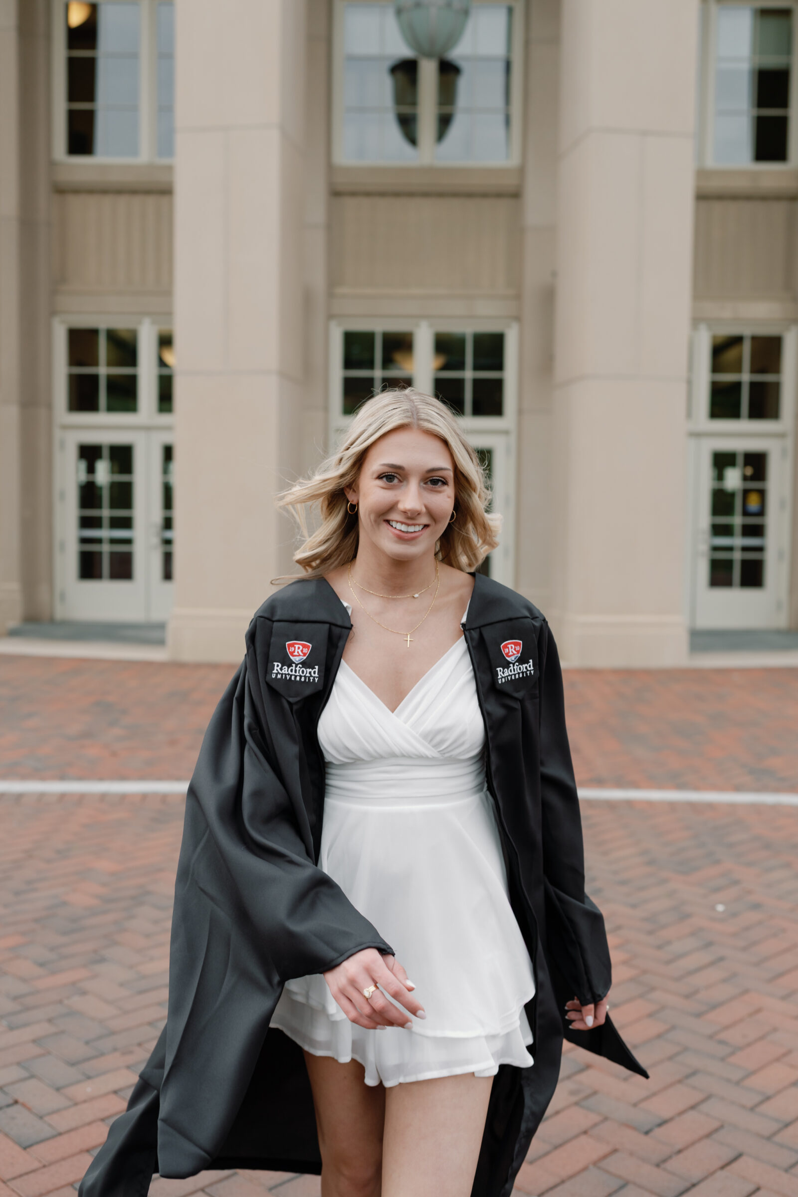 A senior student smiling confidently as she walks toward the camera in front of a university building. She wears a white ruffled dress and her Radford University cap and gown, with her hair flowing and a joyful expression. The brick pathway and columns of the building provide a classic collegiate backdrop.