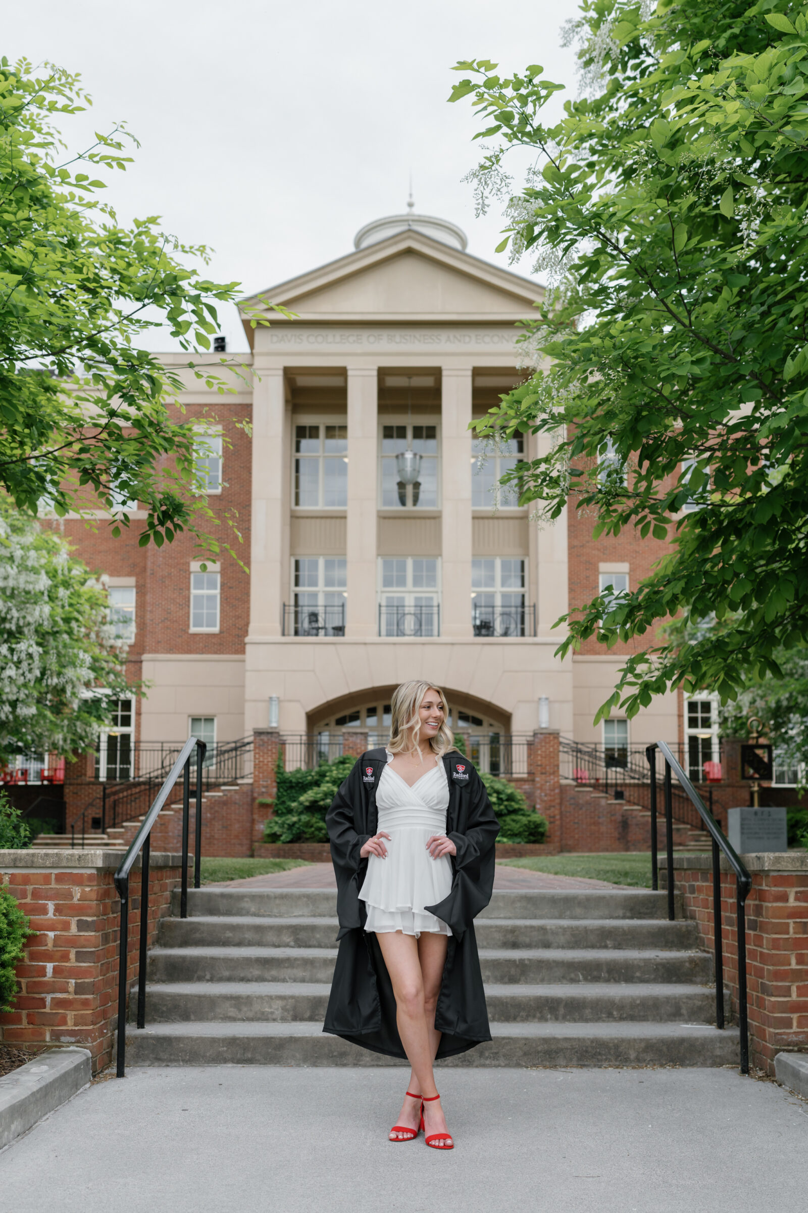 A senior student standing confidently on the steps in front of the Davis College of Business and Economics at Radford University. She wears a white ruffled dress, red heels, and her Radford University cap and gown. Lush greenery frames the image as she smiles, capturing a joyful moment of graduation pride.