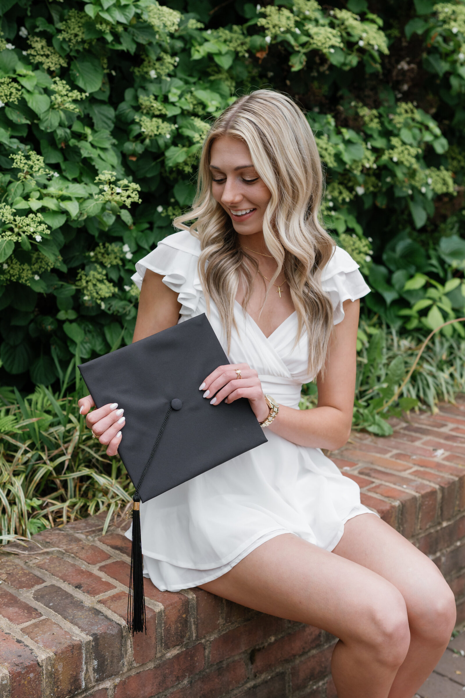 A senior student sitting on a brick wall, holding her graduation cap with a tassel. She smiles down at the cap, wearing a white ruffled dress, a gold watch, and a delicate gold cross necklace, with lush greenery in the background.