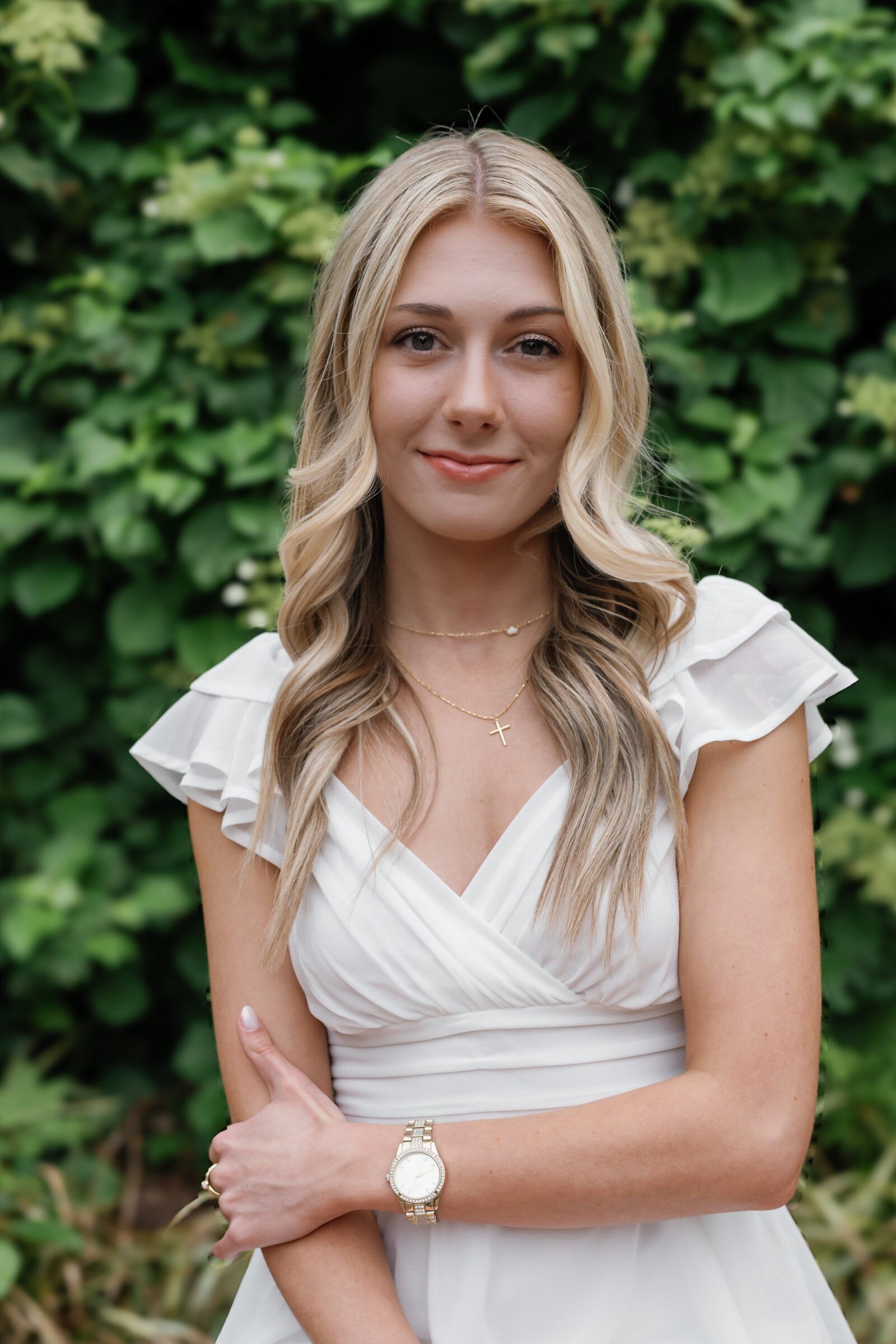 A close-up portrait of a senior student smiling softly in front of lush greenery. She wears a white ruffled dress, layered gold necklaces, and a watch, with her arms crossed elegantly. Her blonde hair is styled in soft waves, complementing her relaxed yet confident pose.