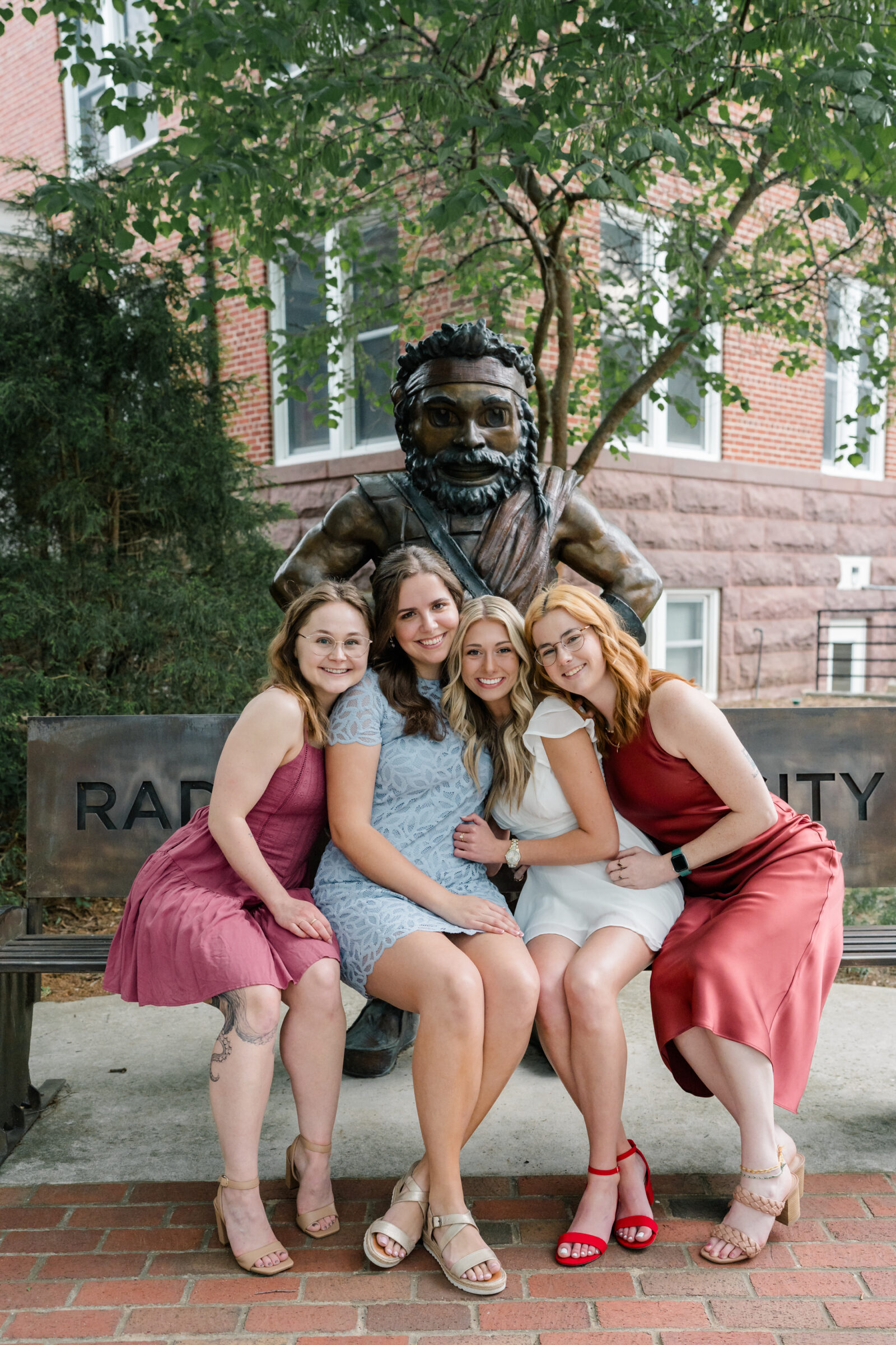 A group of four senior students sitting together on a bench in front of a statue at Radford University. They are dressed in various dresses, with one wearing a white dress. Each student smiles and enjoys the moment, their arms around each other in a joyful, celebratory pose.