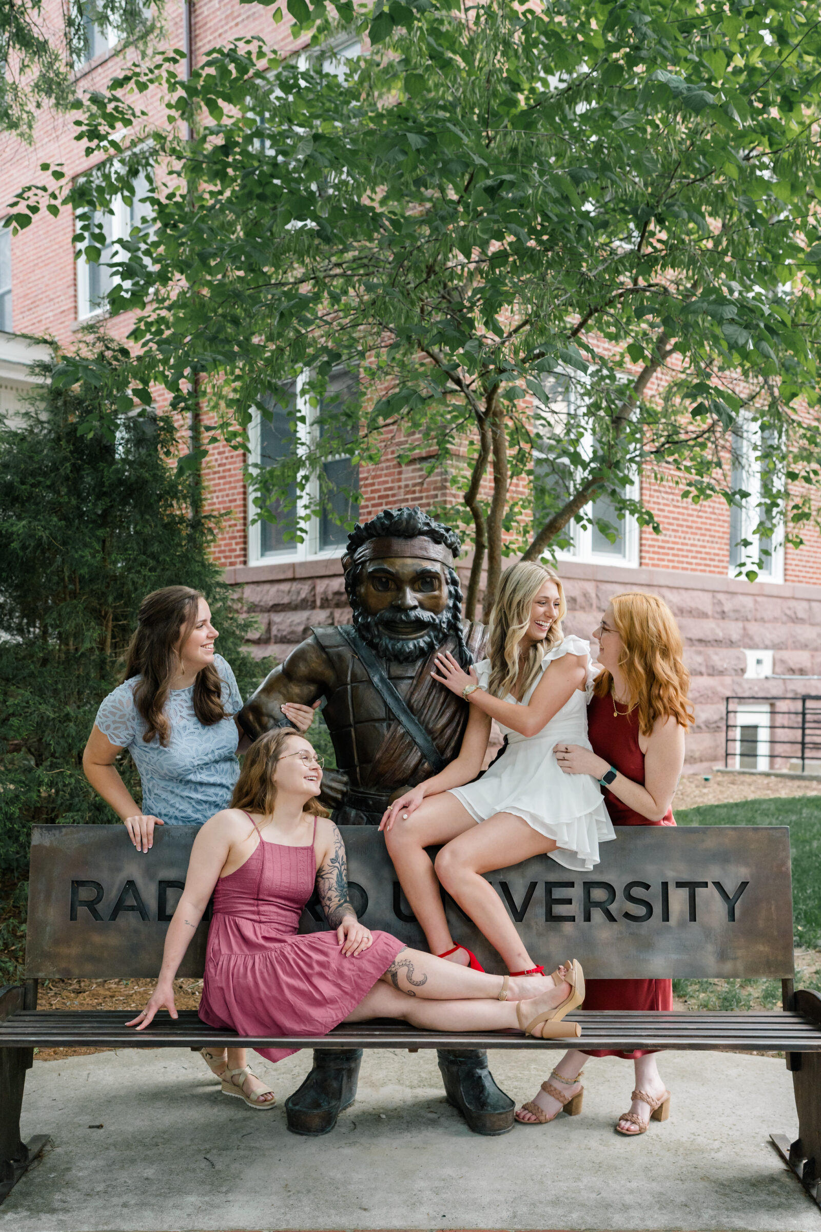 A group of four senior students gathered around a statue at Radford University, laughing and enjoying their time together. They sit on a bench, with one student playfully interacting with the statue. They wear different dresses in various colors, with joyful expressions and relaxed body language.
